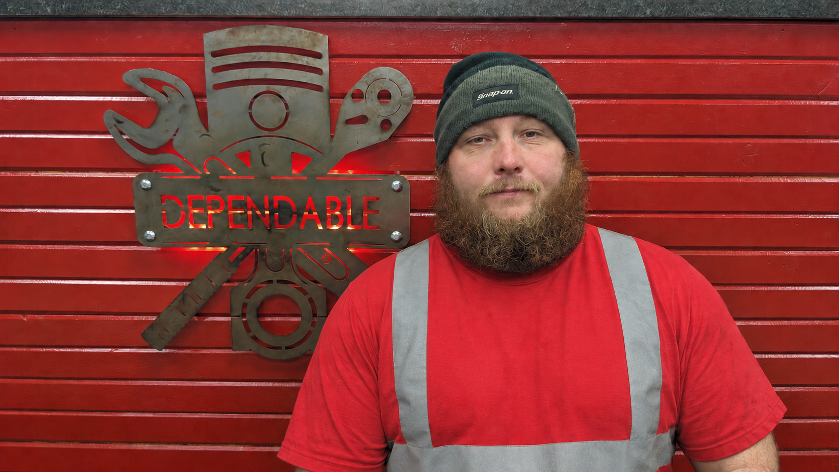 Bearded person in black and red work shirt standing before a red wall with a logo sign. | Dependable Automotive