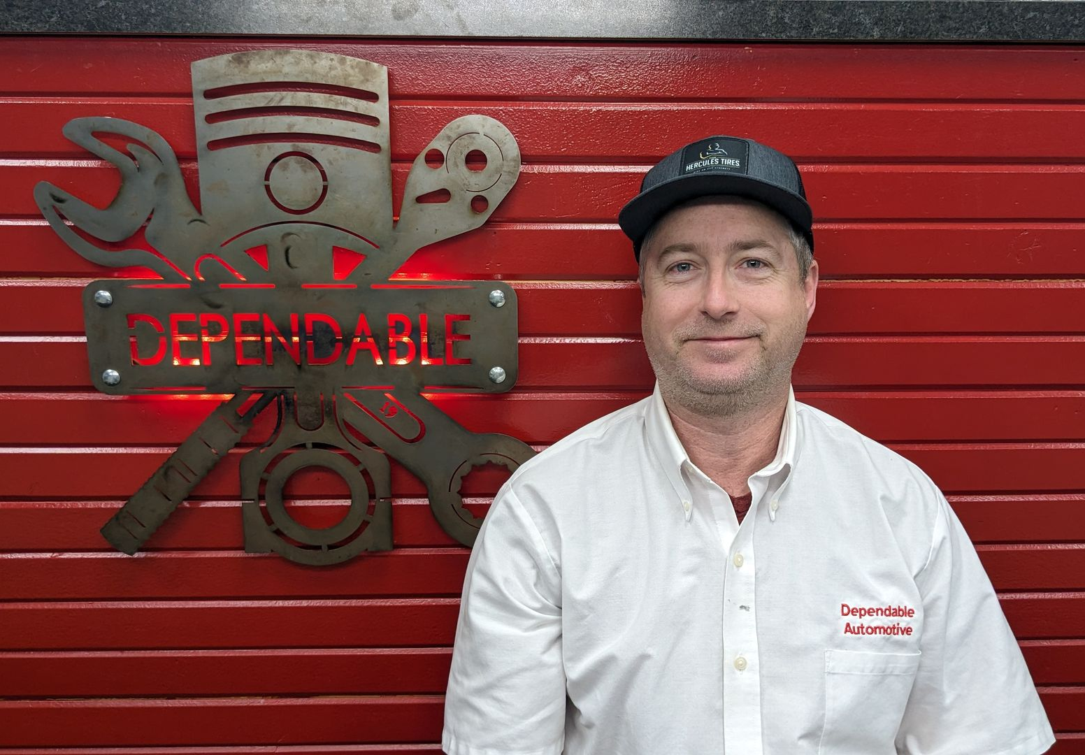 Man in white work shirt and cap standing by red wall with Dependable sign and tools logo | Dependable Automotive