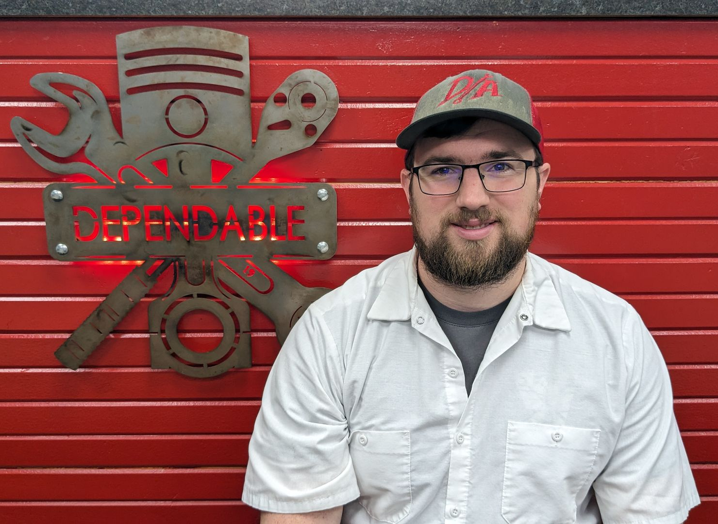 Man in a white shirt and cap stands before a red wall with a “Dependable” mechanic sign. | Dependable Automotive