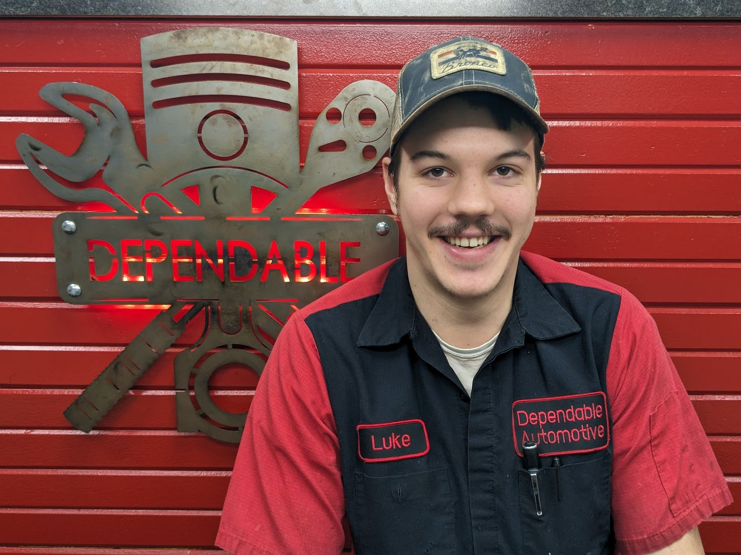 Smiling mechanic in red-and-black uniform and cap standing before a red Dependable sign. | Dependable Automotive