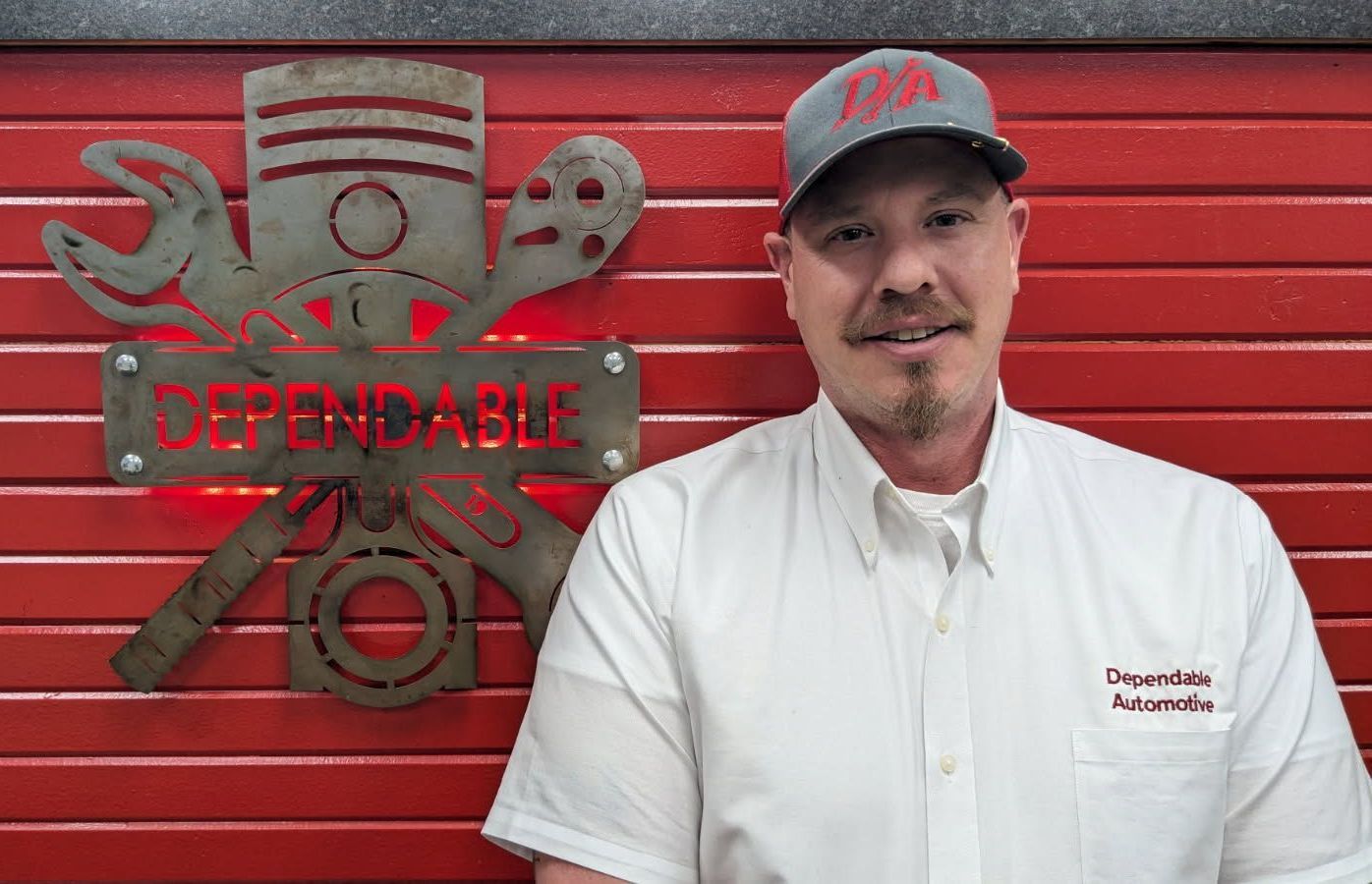 Man standing by a red garage wall with a large “Dependable” tool logo, wearing a white shirt and gray cap | Dependable Automotive