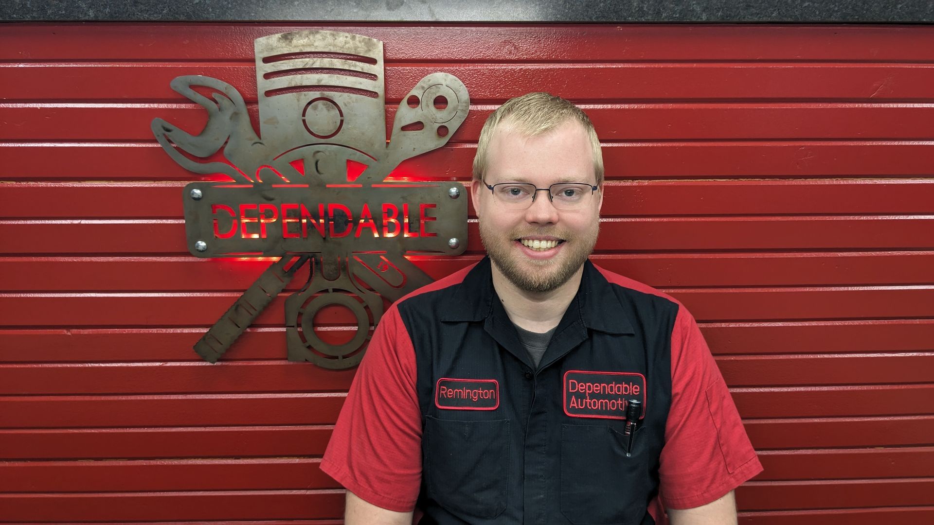 Man smiling in black and red work shirt standing before a red garage door and shop logo. | Dependable Automotive