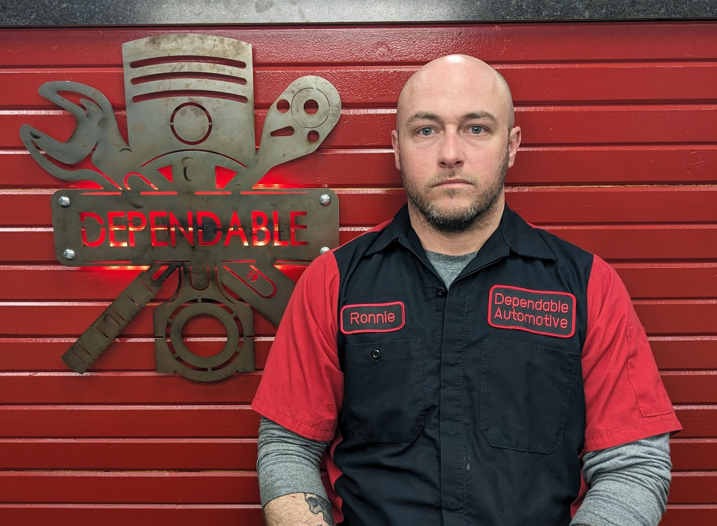 Man in black and red work shirt standing beside a Dependable automotive logo on a red wall | Dependable Automotive