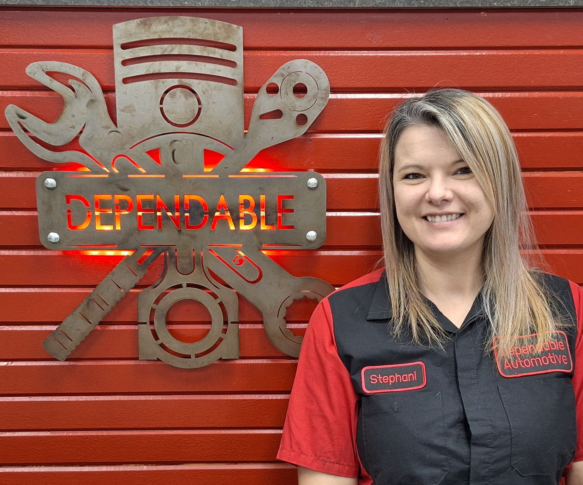 Woman in a red-and-black uniform smiling beside a red wall with a metal “DEPENDABLE” emblem. | Dependable Automotive