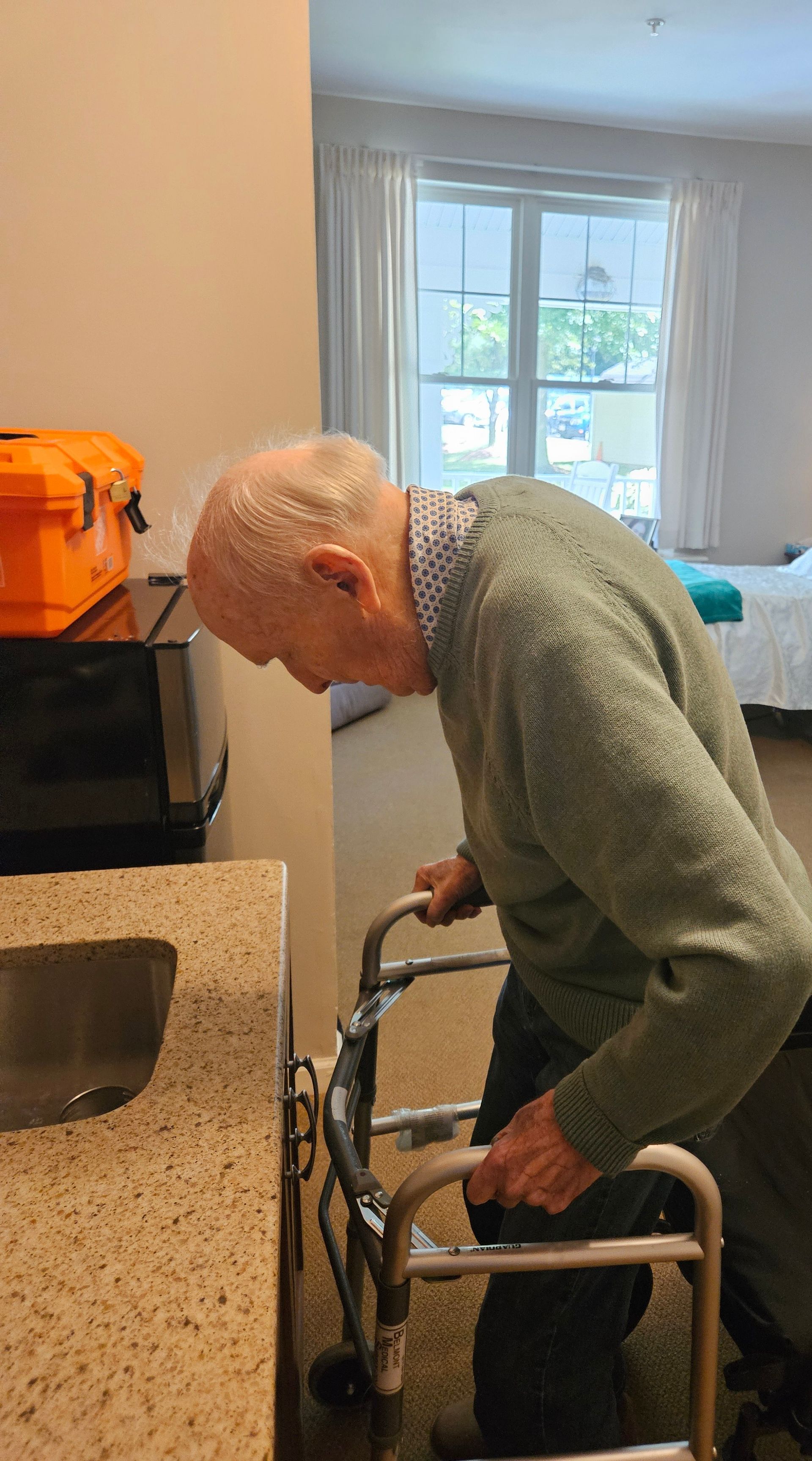 An elderly person uses a walker near a sink in a room with a window, leaning forward with a light green sweater.
