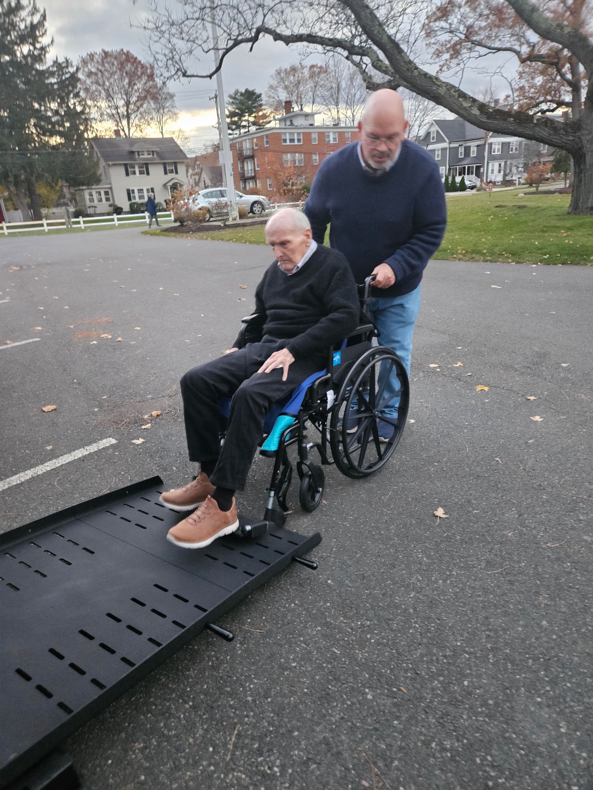Man in wheelchair being pushed up a ramp by another person on a paved area, with park and buildings in background.
