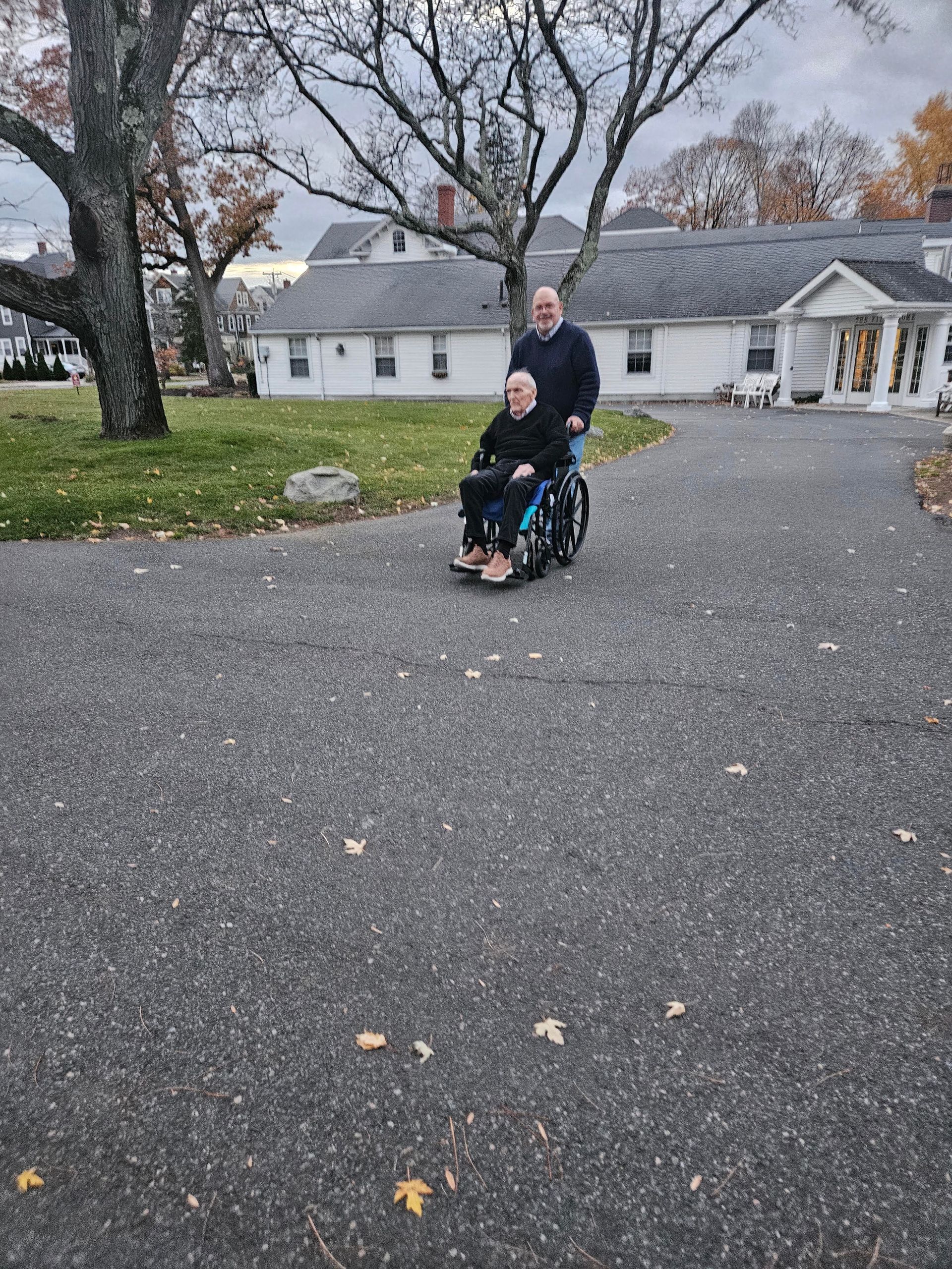 Person in wheelchair being pushed on a driveway by another person, building in background.