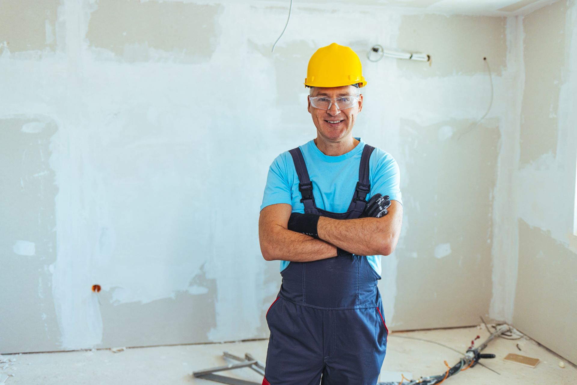 A man wearing a hard hat and overalls is standing in a room with his arms crossed.