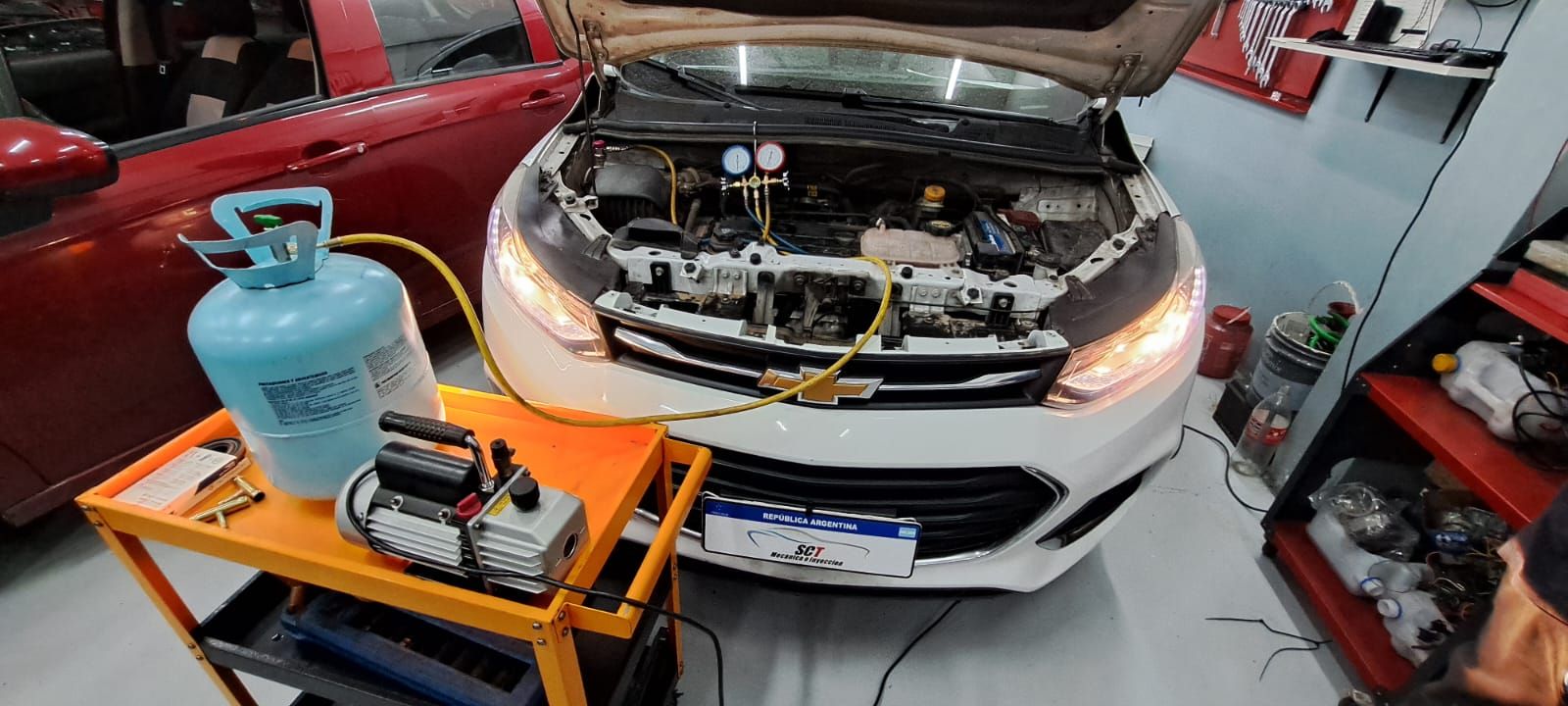 Un automóvil blanco con el capó abierto, recibiendo servicio en un taller con equipo de aire acondicionado.