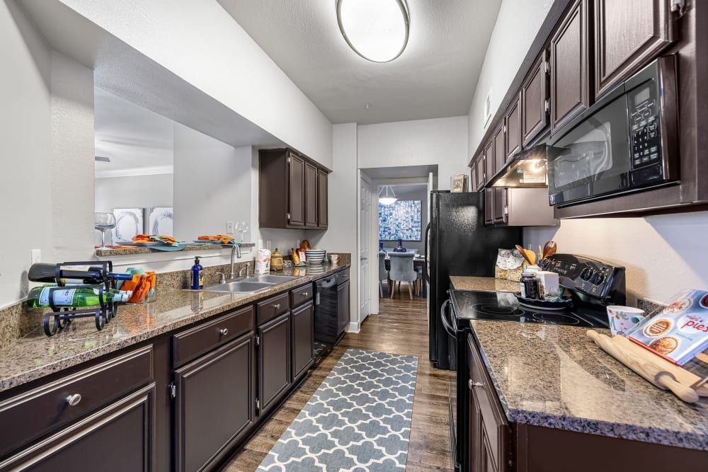a kitchen with brown cabinets, granite counter tops, black appliances, and a rug at Marquis on Pin Oak in Houston, TX.