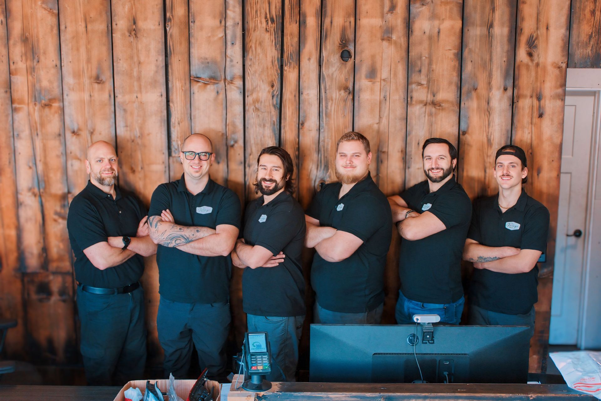 Six men in black shirts standing in front of a wooden wall, arms crossed.