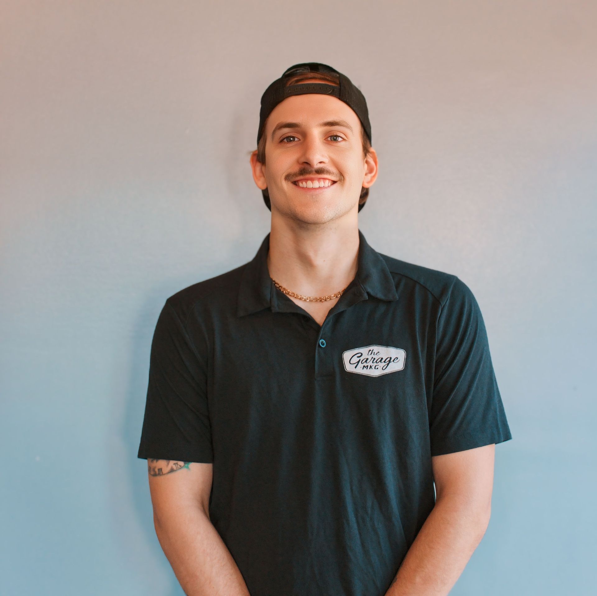 Man with mustache smiles, wearing a black polo shirt and hat against a blue-pink gradient background.