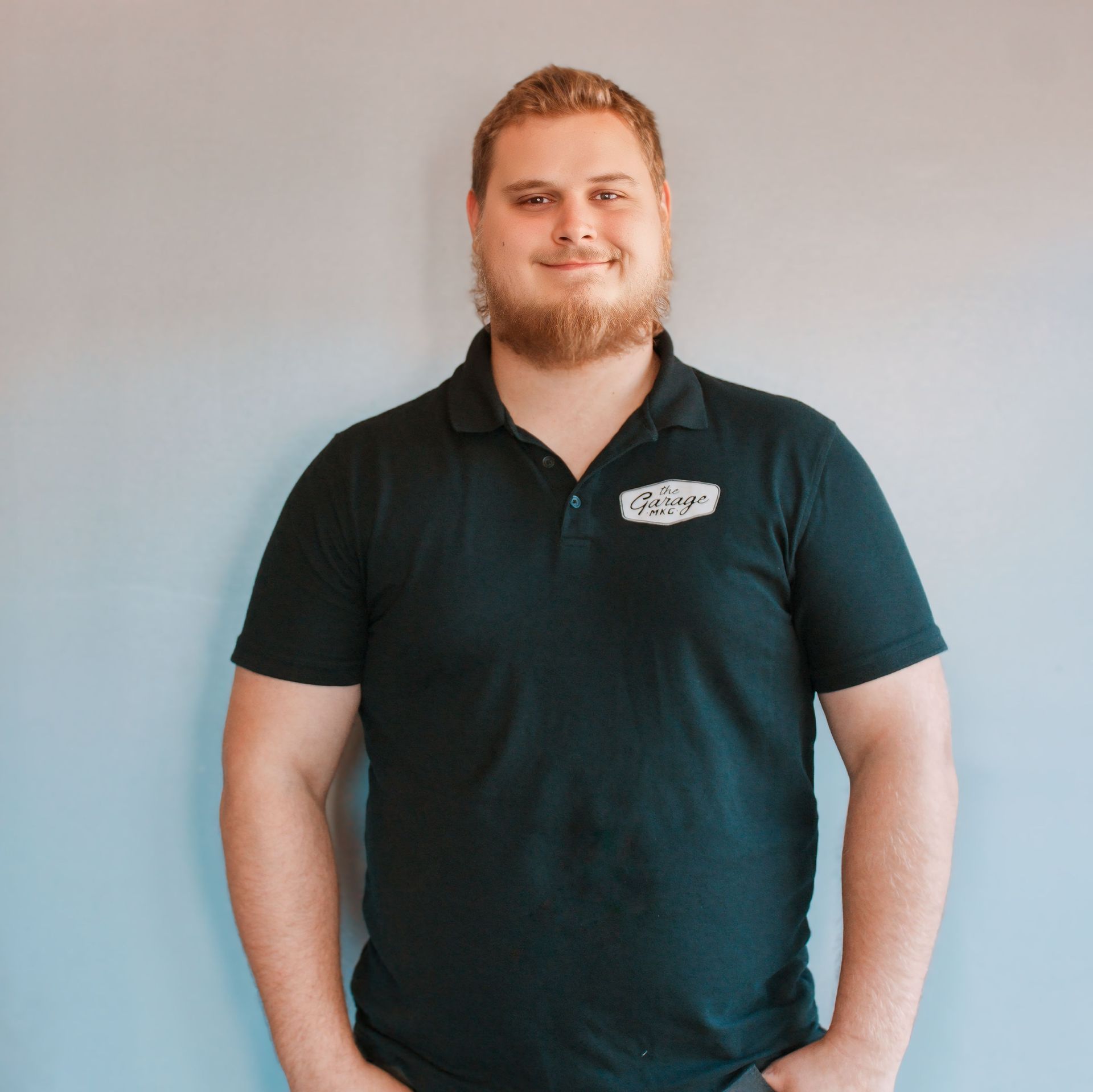 Man with blonde hair and beard in a dark green polo shirt stands against a grey-blue wall; smiling.