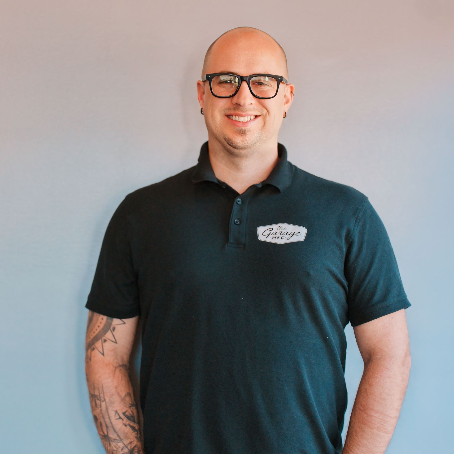Man with glasses and shaved head wearing a dark green polo shirt smiles. Tattoo visible on left arm.
