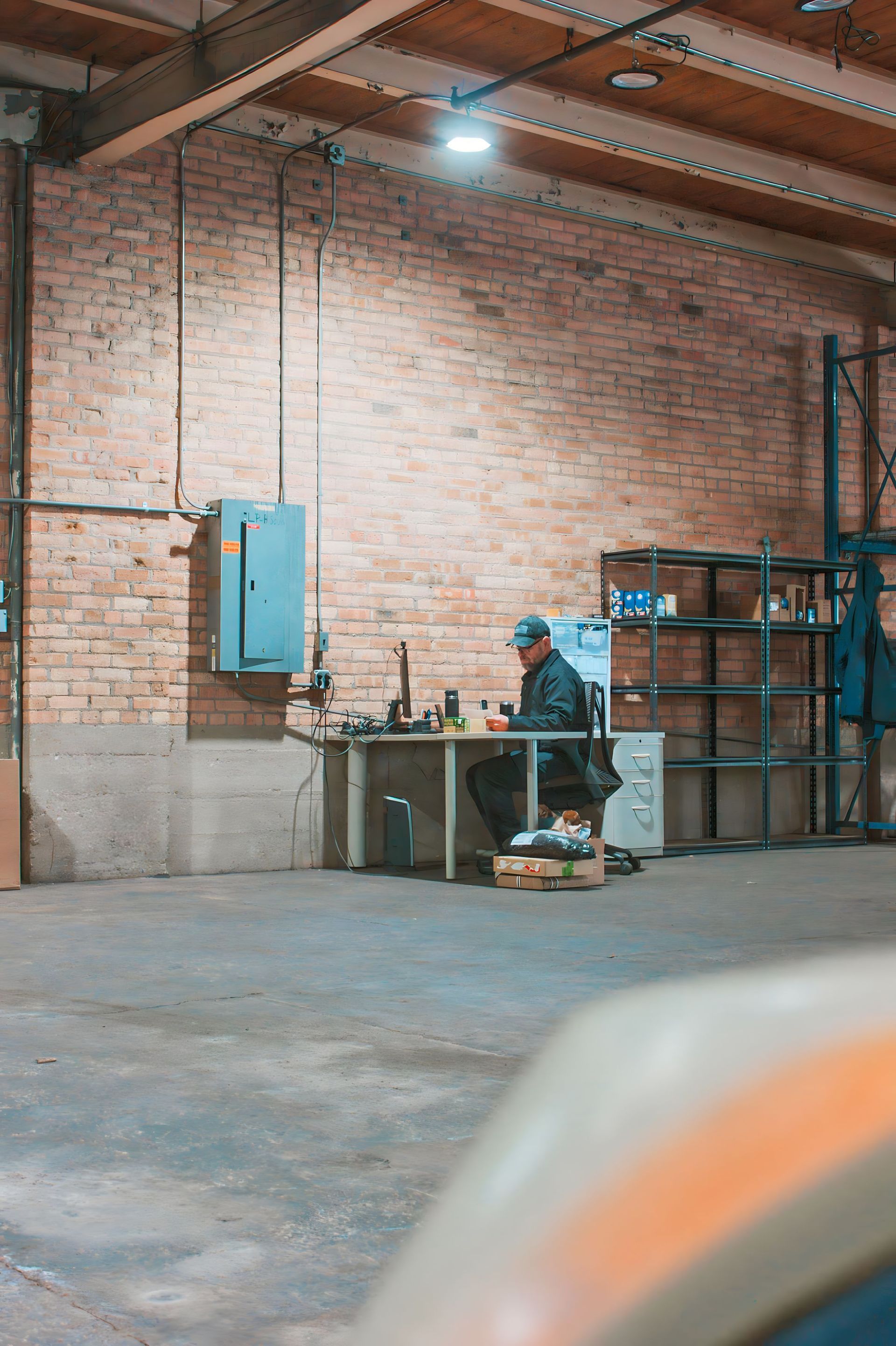 Person working at a desk in a brick-walled warehouse. They are seated and surrounded by equipment.