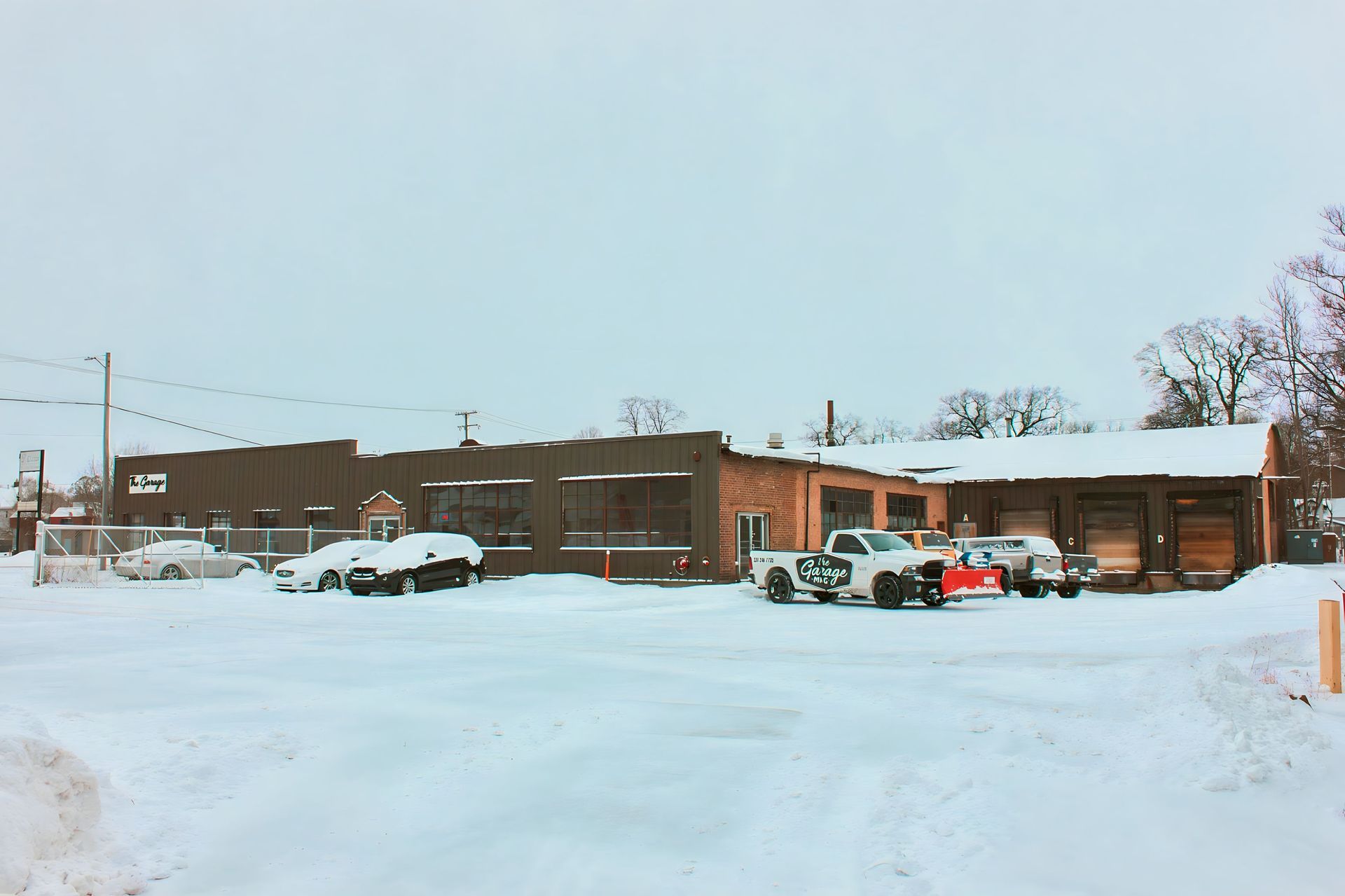 Snow-covered industrial building with parked cars and trucks in a snowy parking lot under a grey sky.
