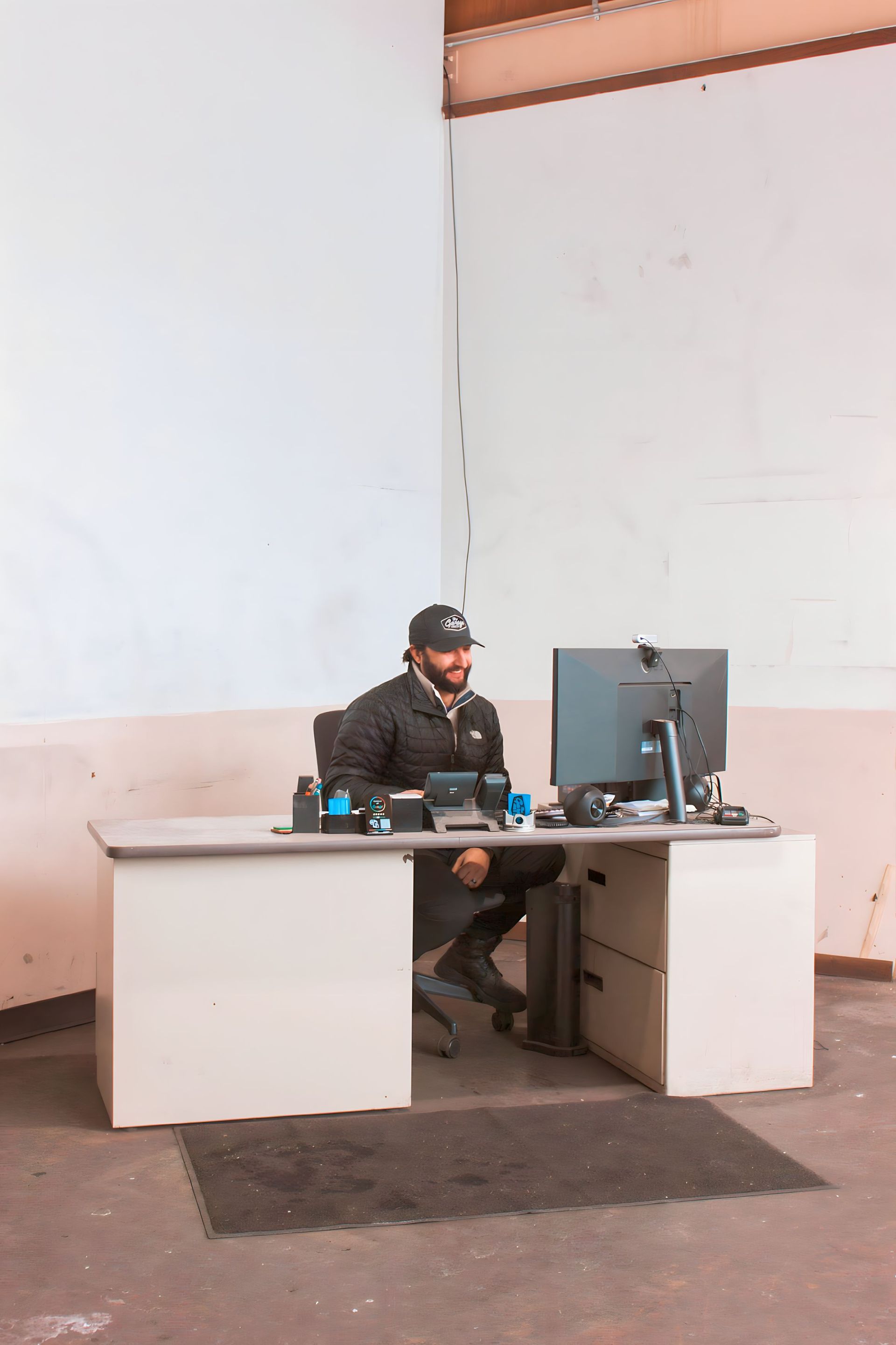Man at a desk in an empty warehouse, looking at a computer. He's wearing a cap and jacket.