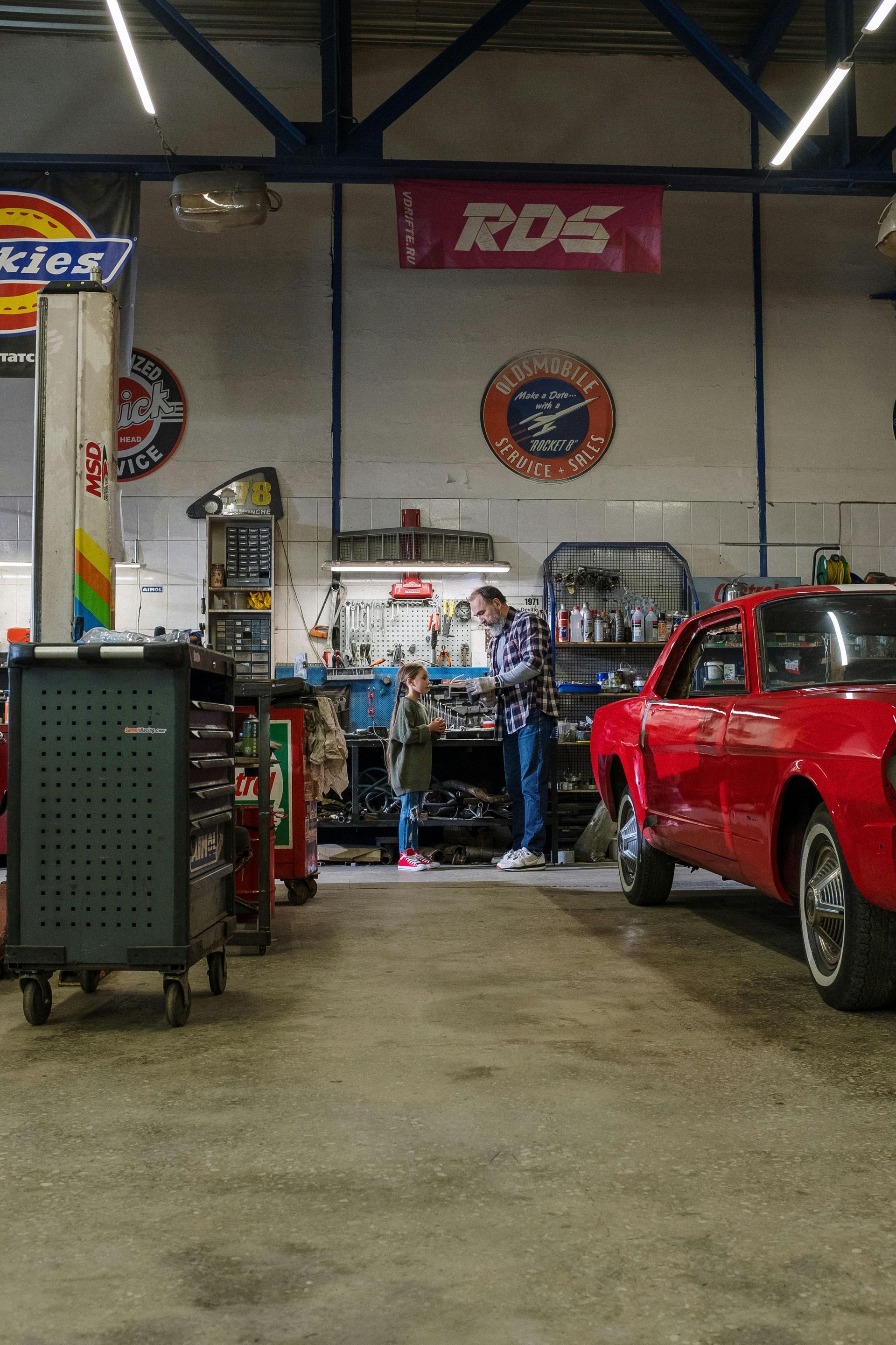 Man and child in a garage, working on a car. Red car parked nearby.