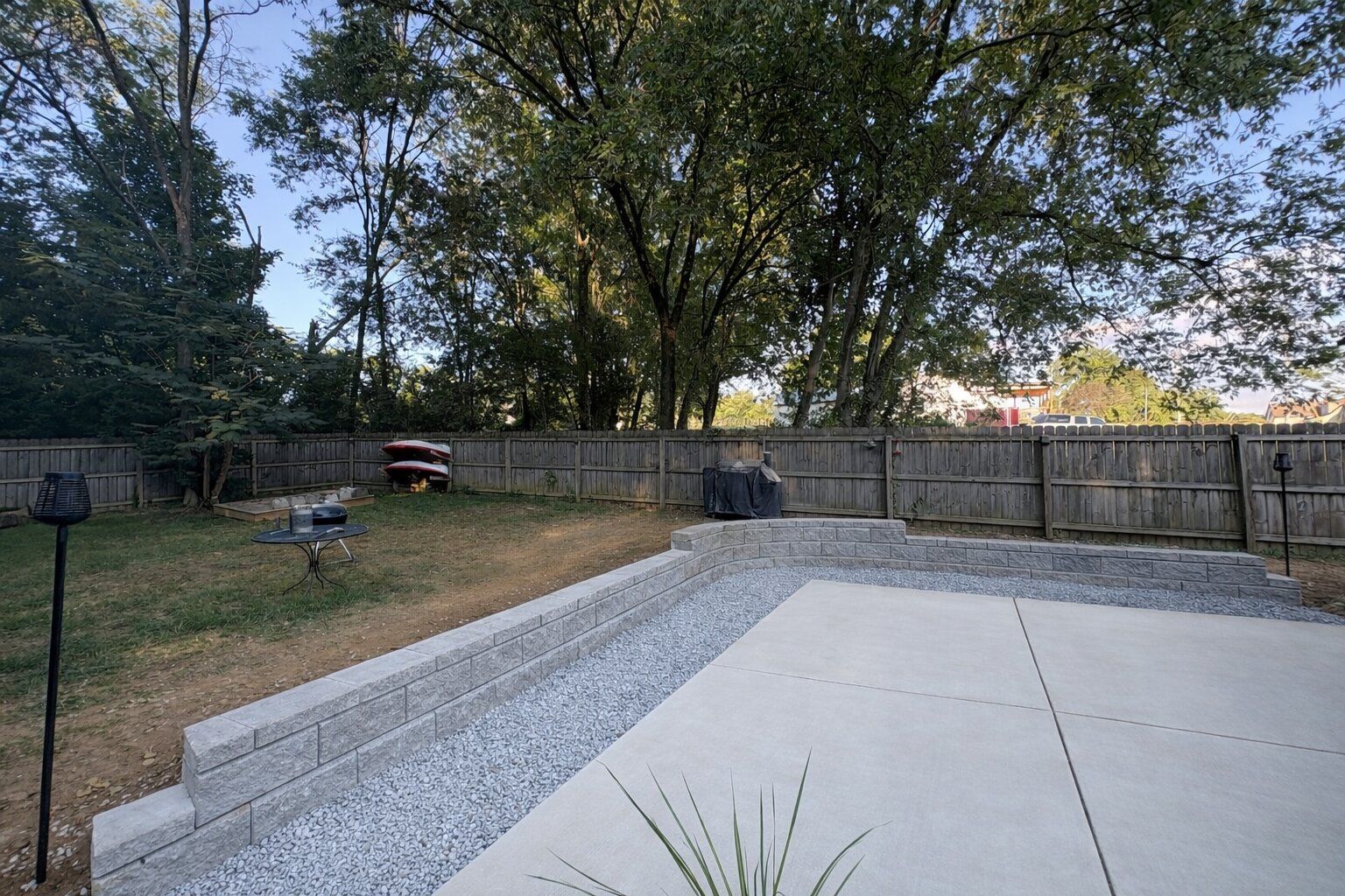 A concrete patio with a stone retaining wall borders a grassy backyard, framed by a wooden fence and tall trees.