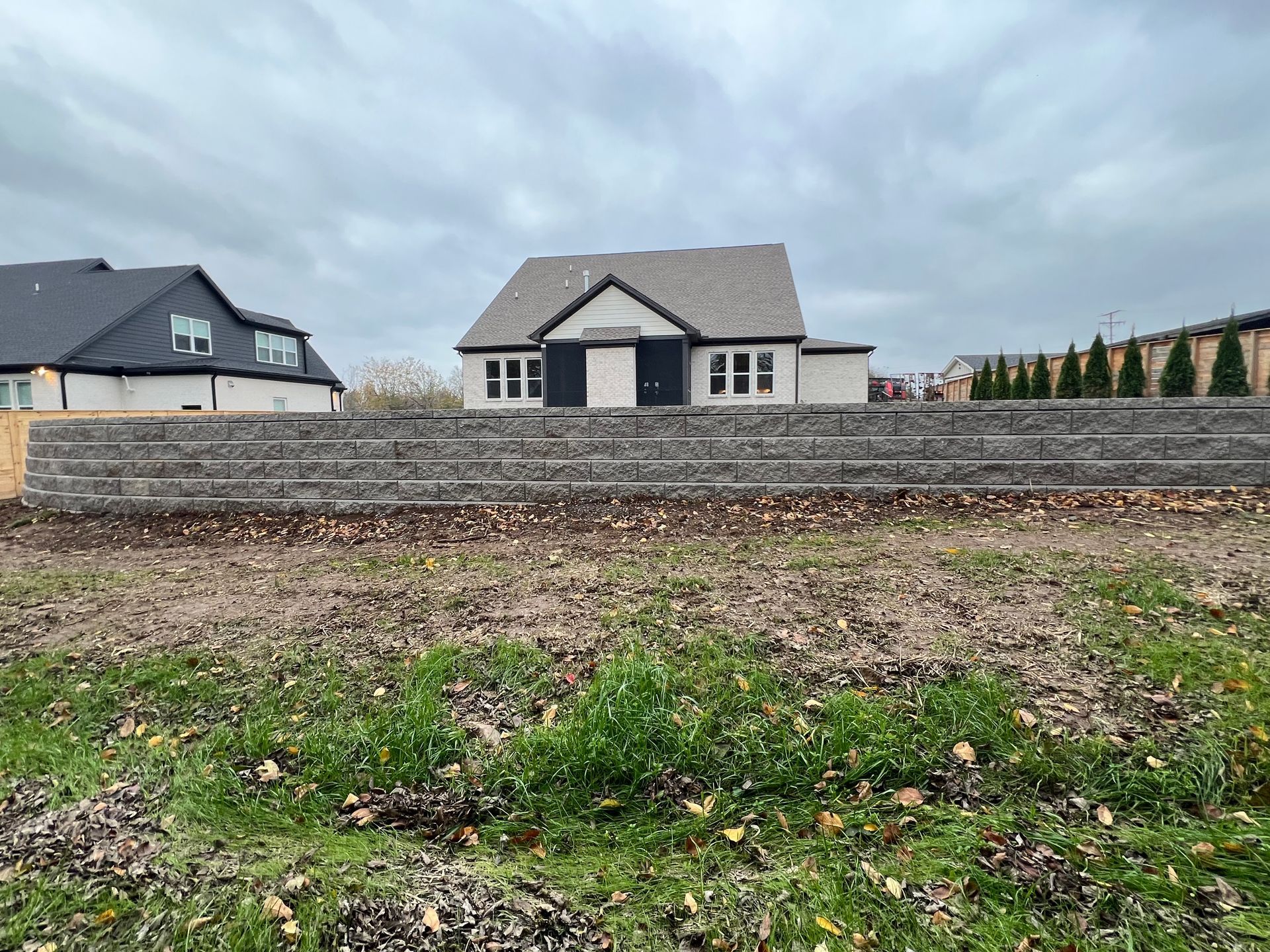 A modern house sits behind a large stone retaining wall on a cloudy day, with a neighboring house on the left.