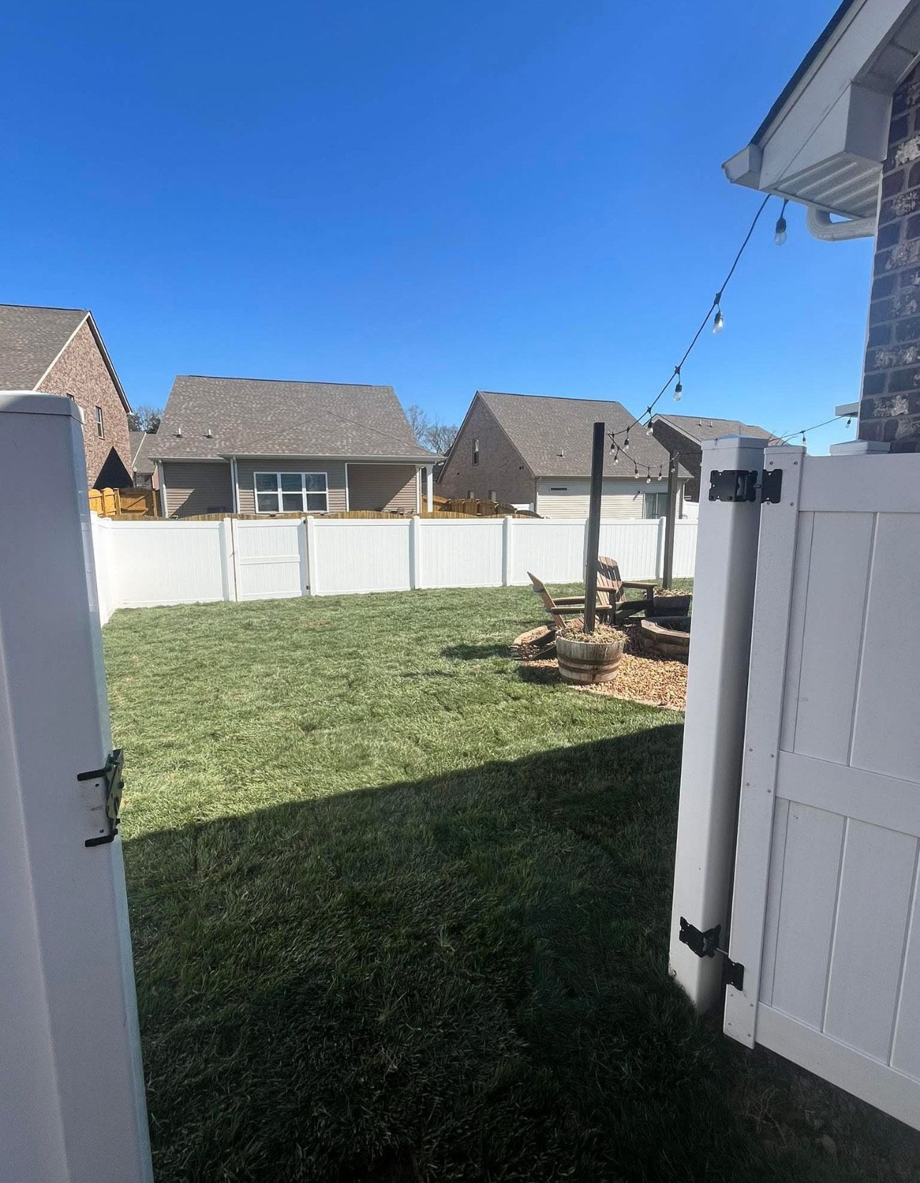 Backyard with mulch, patio furniture, retaining wall, trees, and houses under a blue sky.