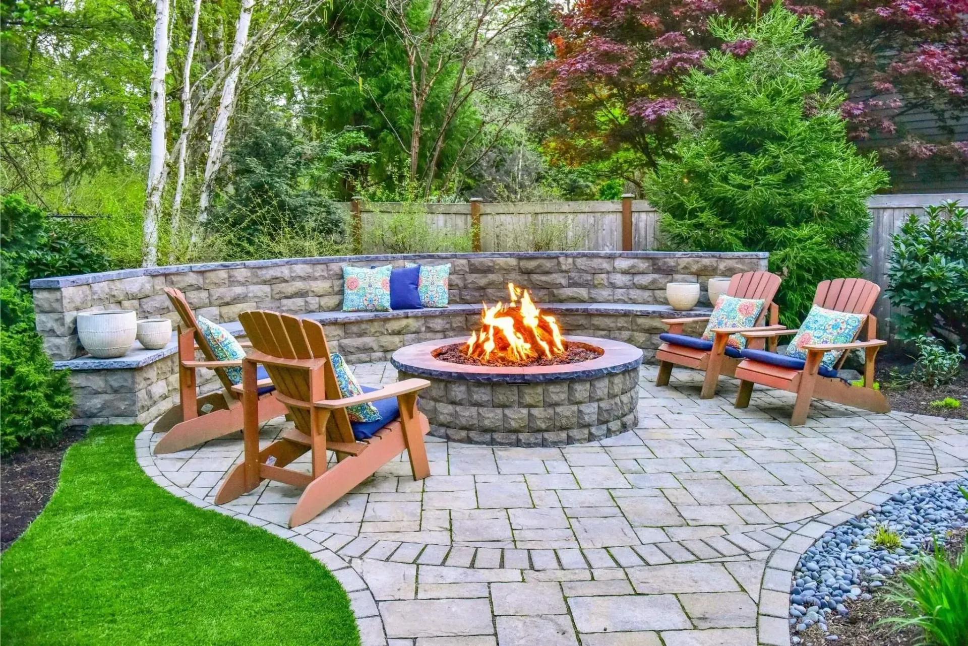 A circular stone fire pit with a bright fire burns in a stone-paved patio, surrounded by four wooden chairs in a garden.