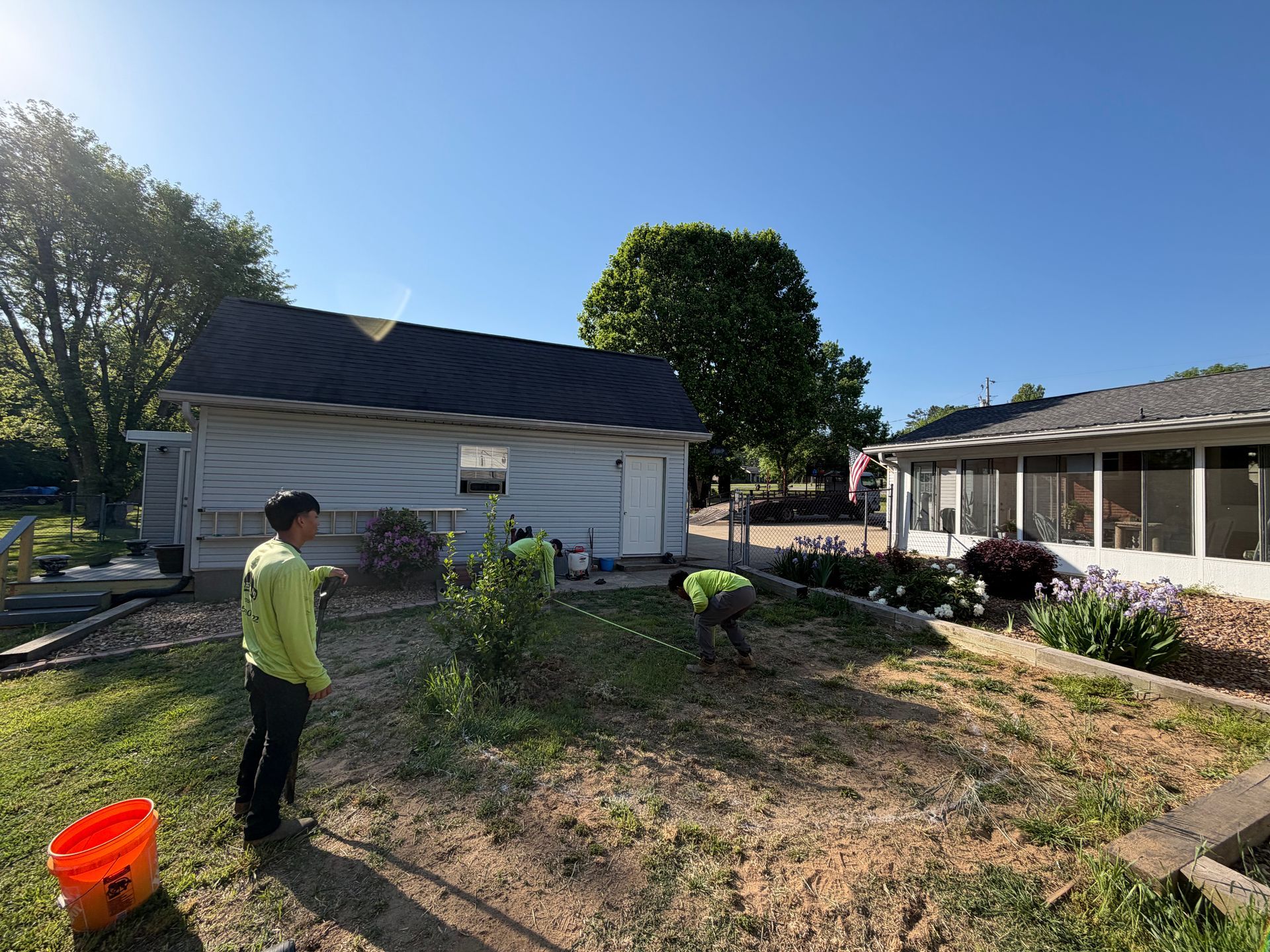 People working in a sunny backyard beside a white house and garden beds