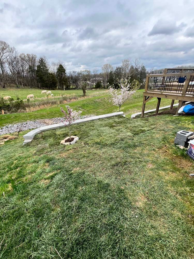 Green yard with a concrete border, fire pit, and a wooden deck under a cloudy sky.