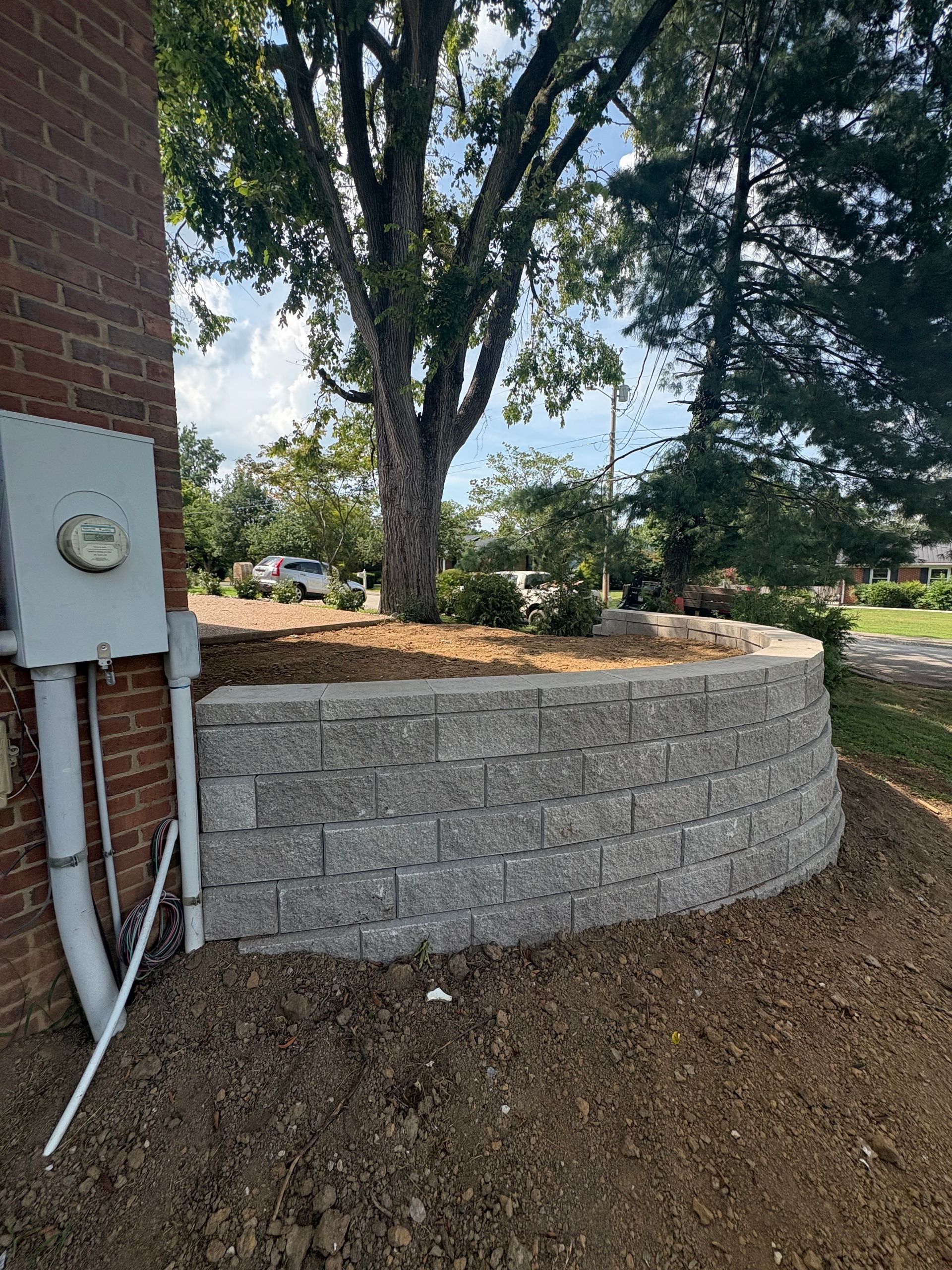 Gray stone retaining wall curves around a tree on a sloped yard, next to a brick building.