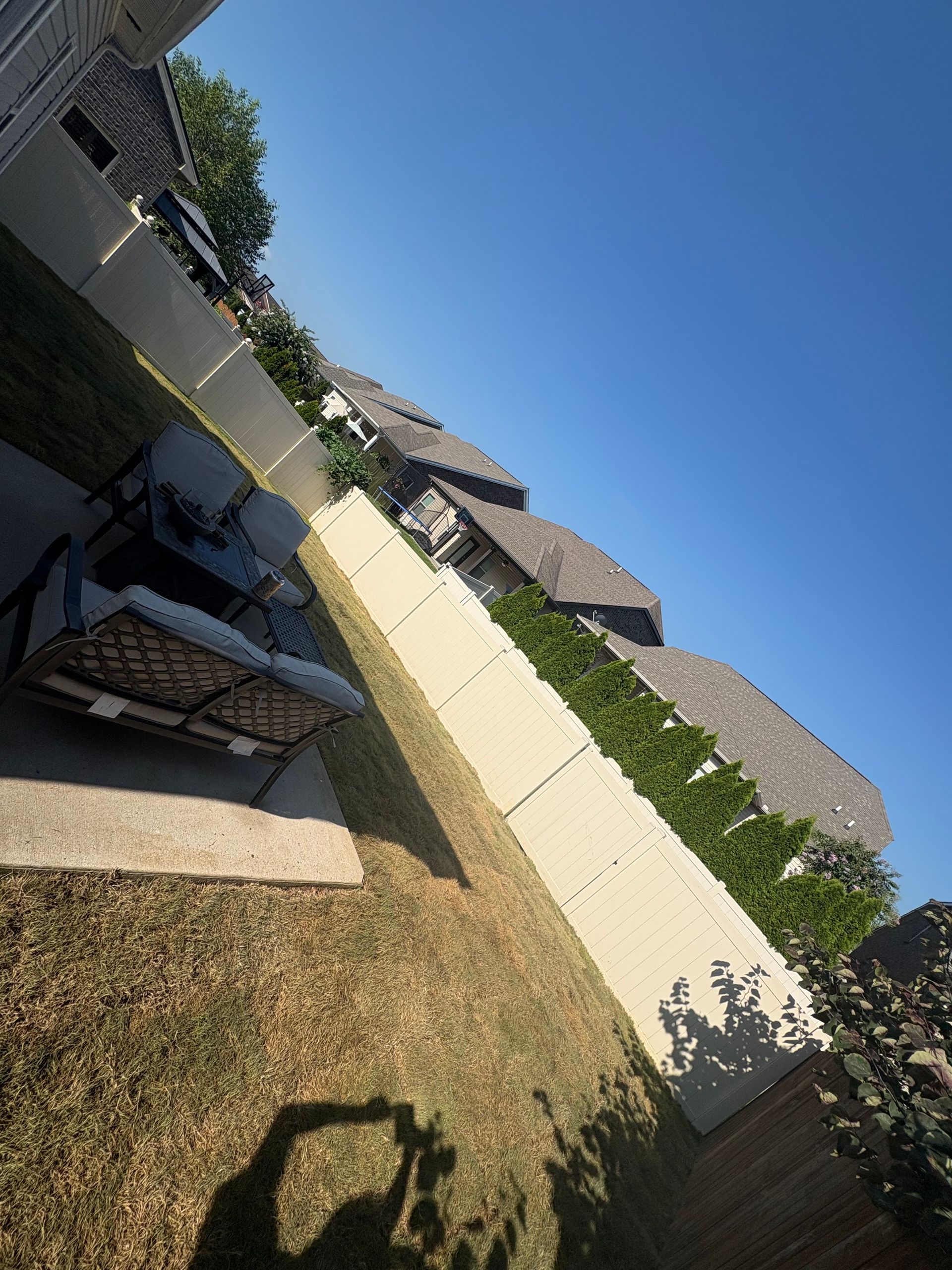 Backyard with mulch, patio furniture, retaining wall, trees, and houses under a blue sky.