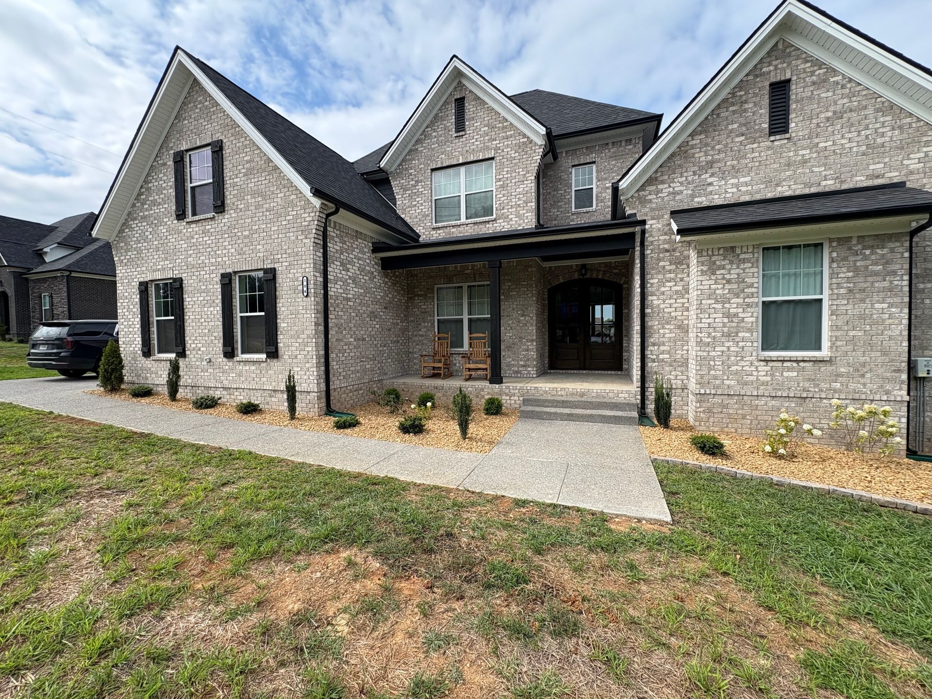 Gray brick house with black accents, front porch, and landscaping under a cloudy sky.