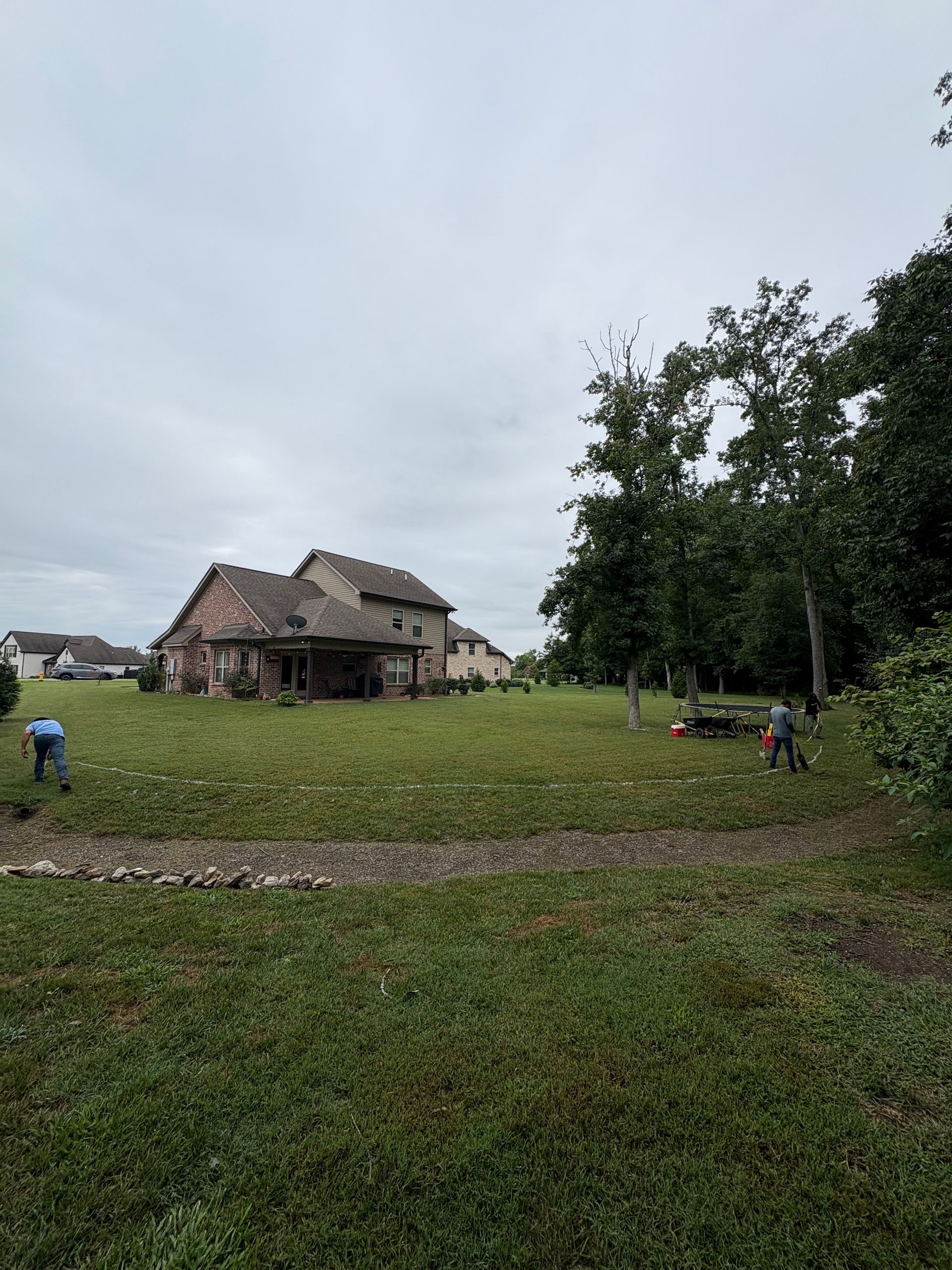 People work on a grassy field in front of a weathered building under a cloudy sky.