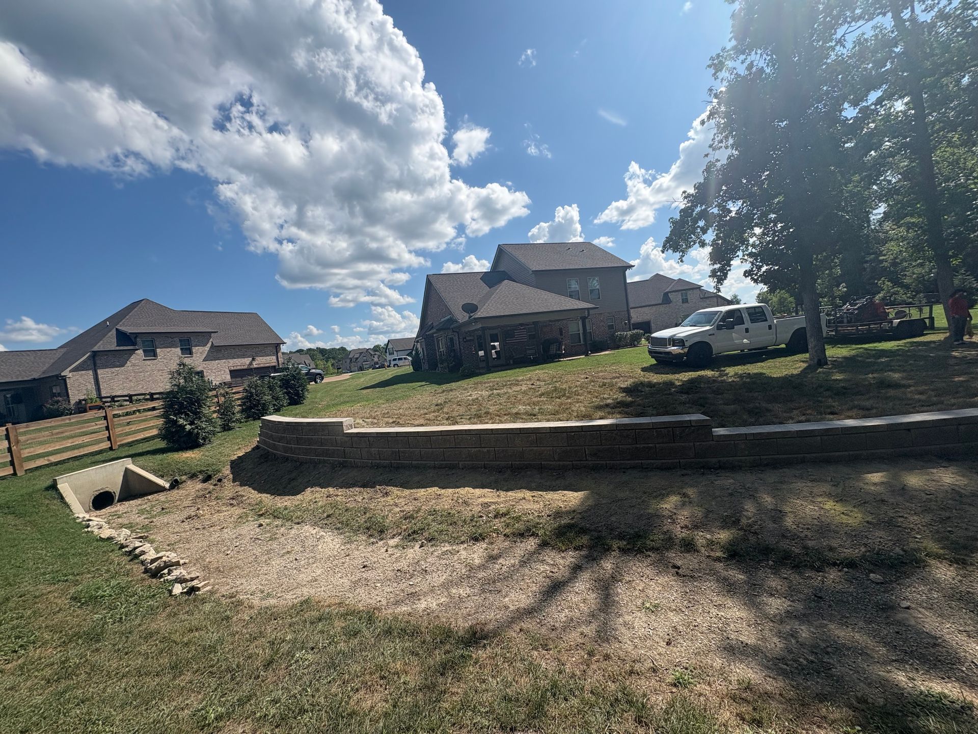 A dry creek bed in a residential area with houses, a retaining wall, and a white truck. Sunny, blue sky.