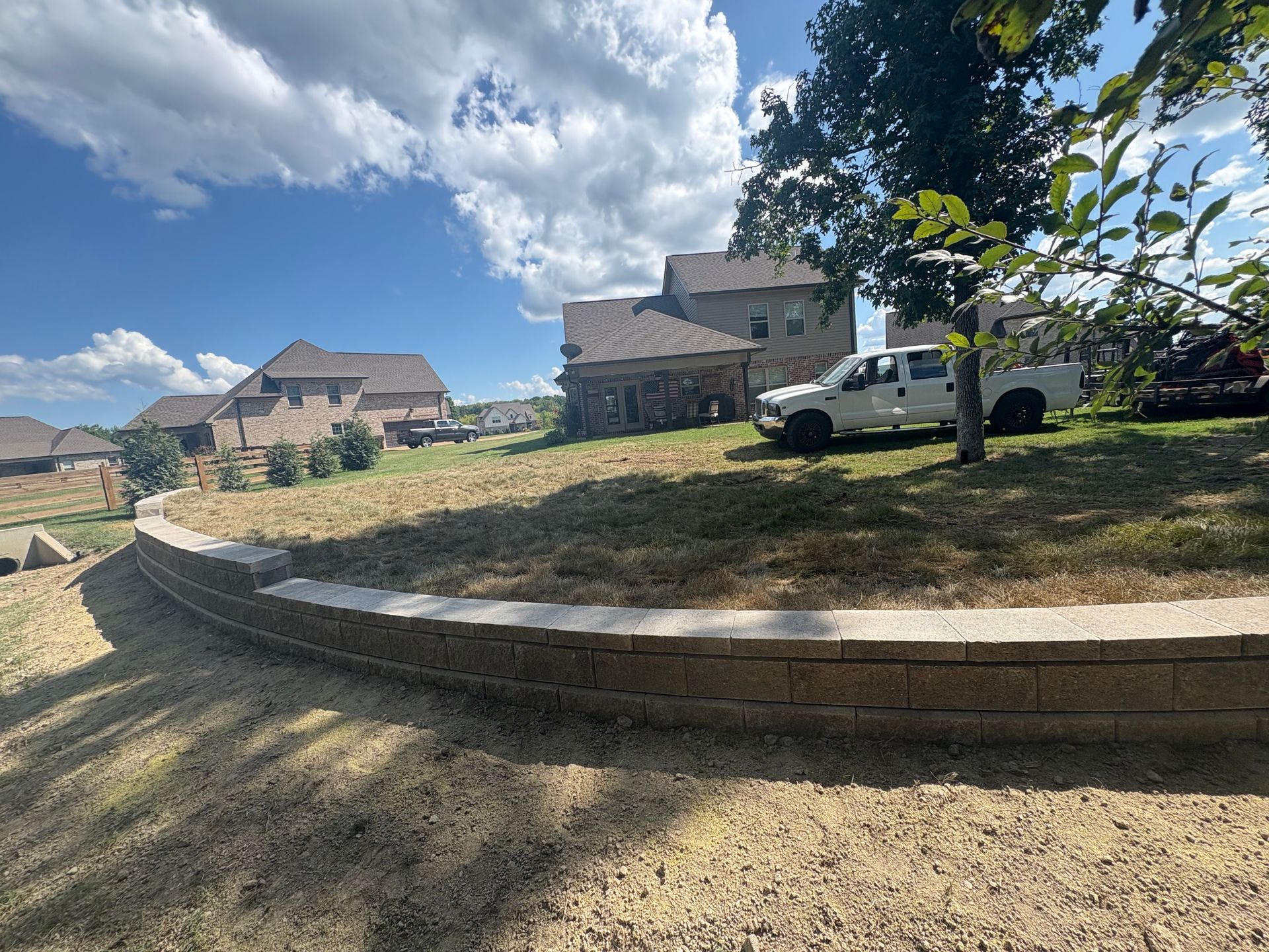 A retaining wall with a truck parked on a grassy hill, houses in the distance under a cloudy sky.