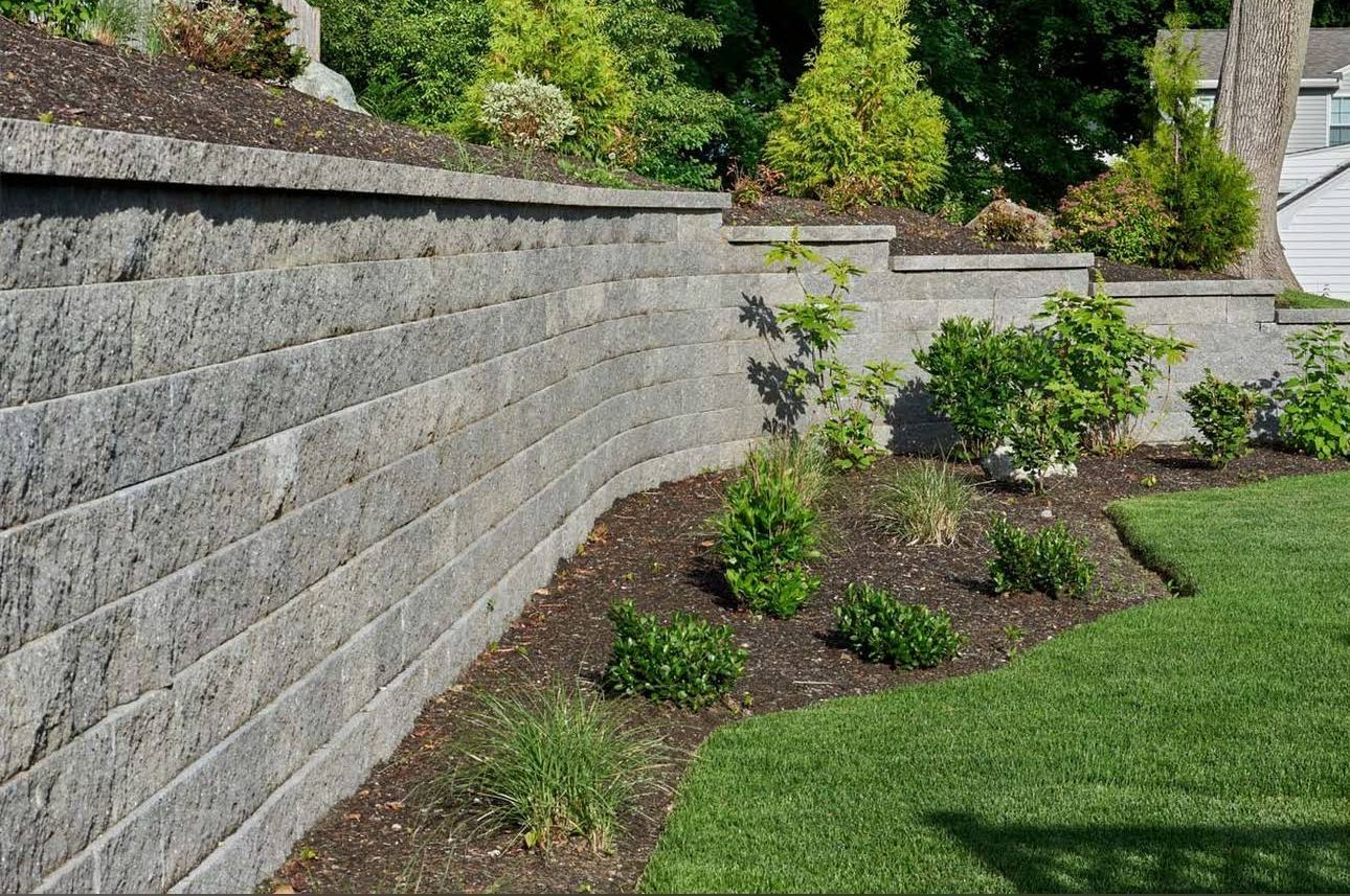 A tiered grey stone retaining wall borders a garden bed with small shrubs, mulch, and a green lawn.
