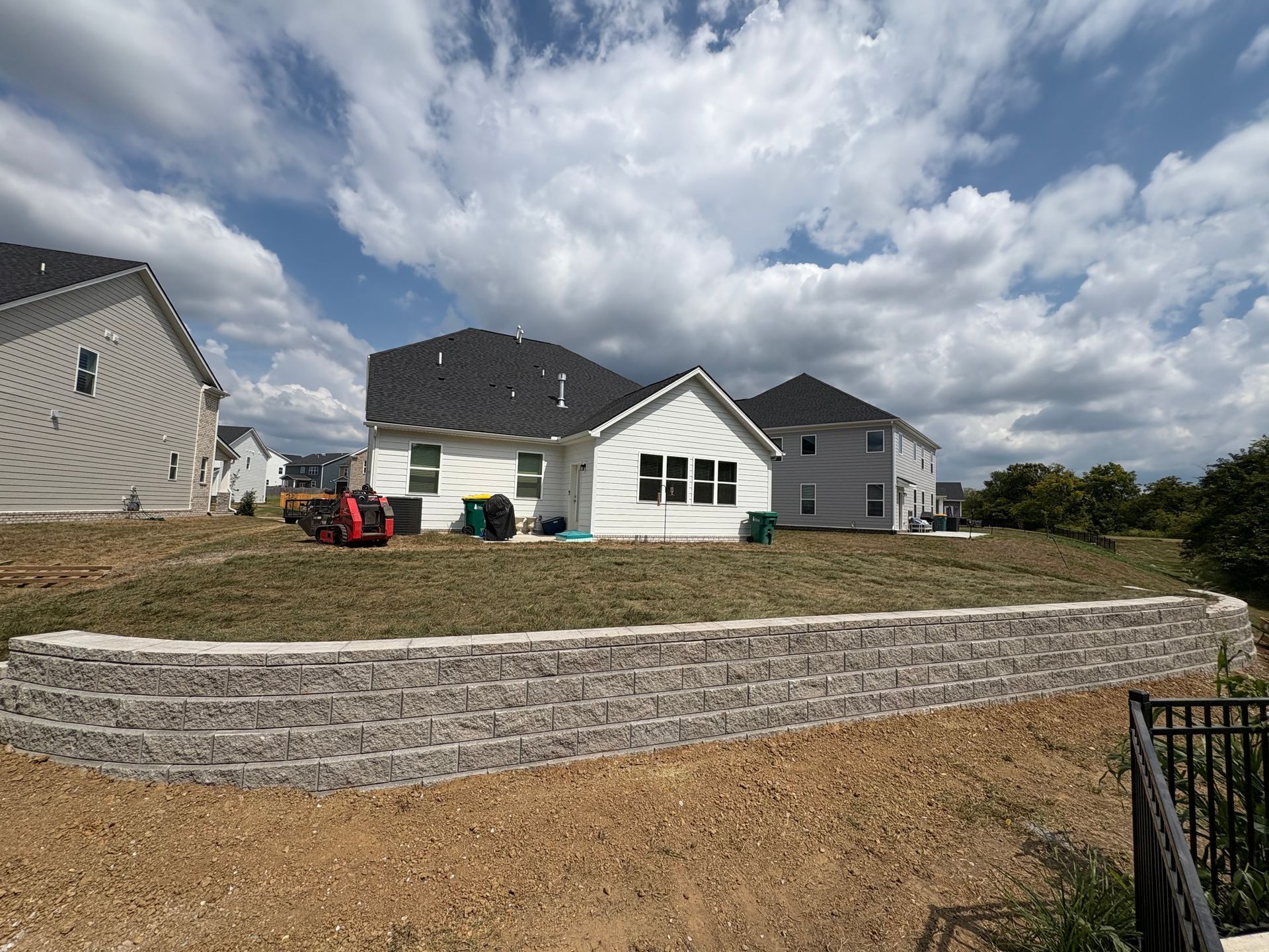A grey stone retaining wall in front of a white house with a dark shingled roof under a partly cloudy sky.