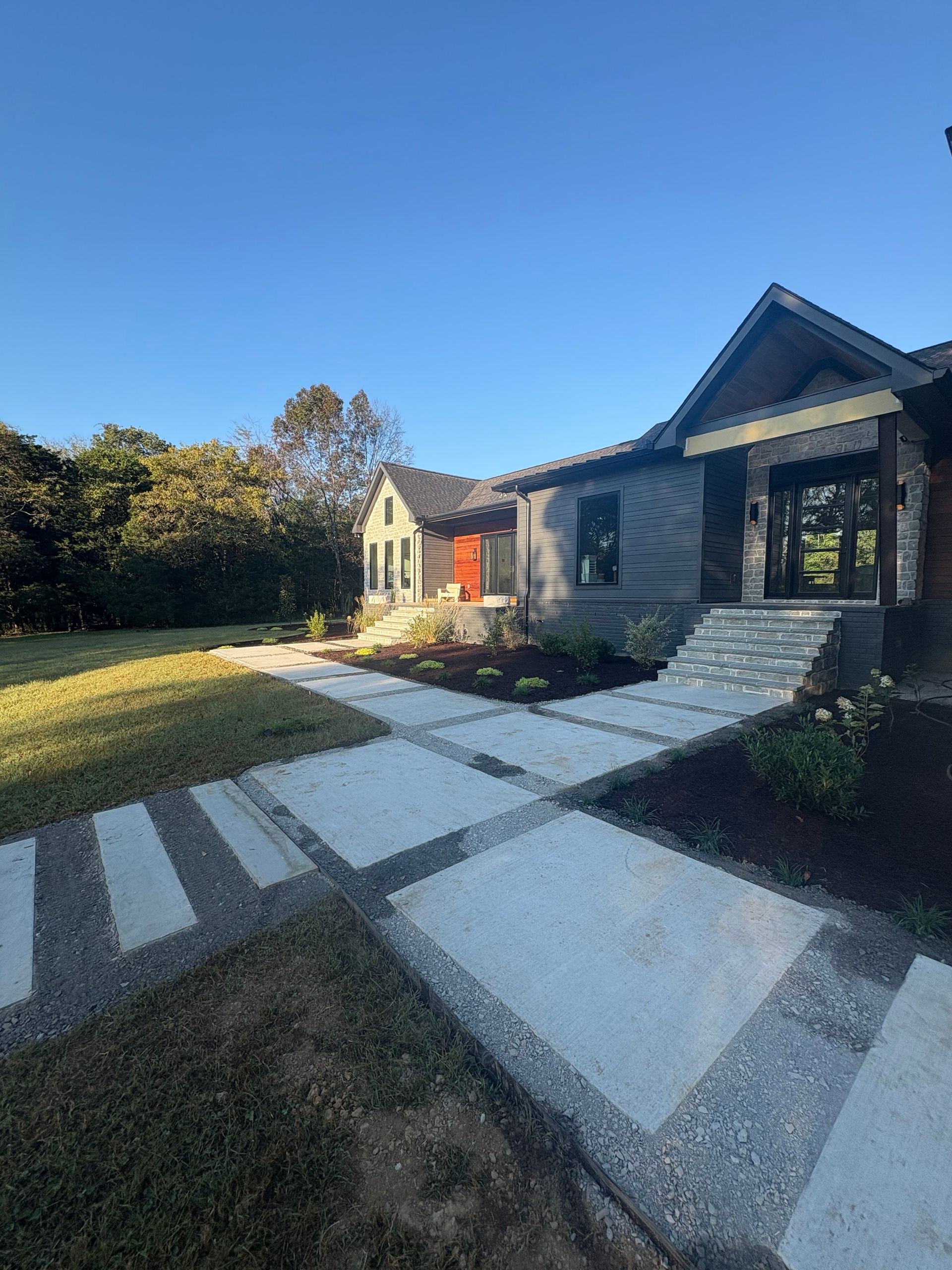 A modern house exterior with a gray facade, orange door, and a geometric concrete paver walkway leading to the entrance.