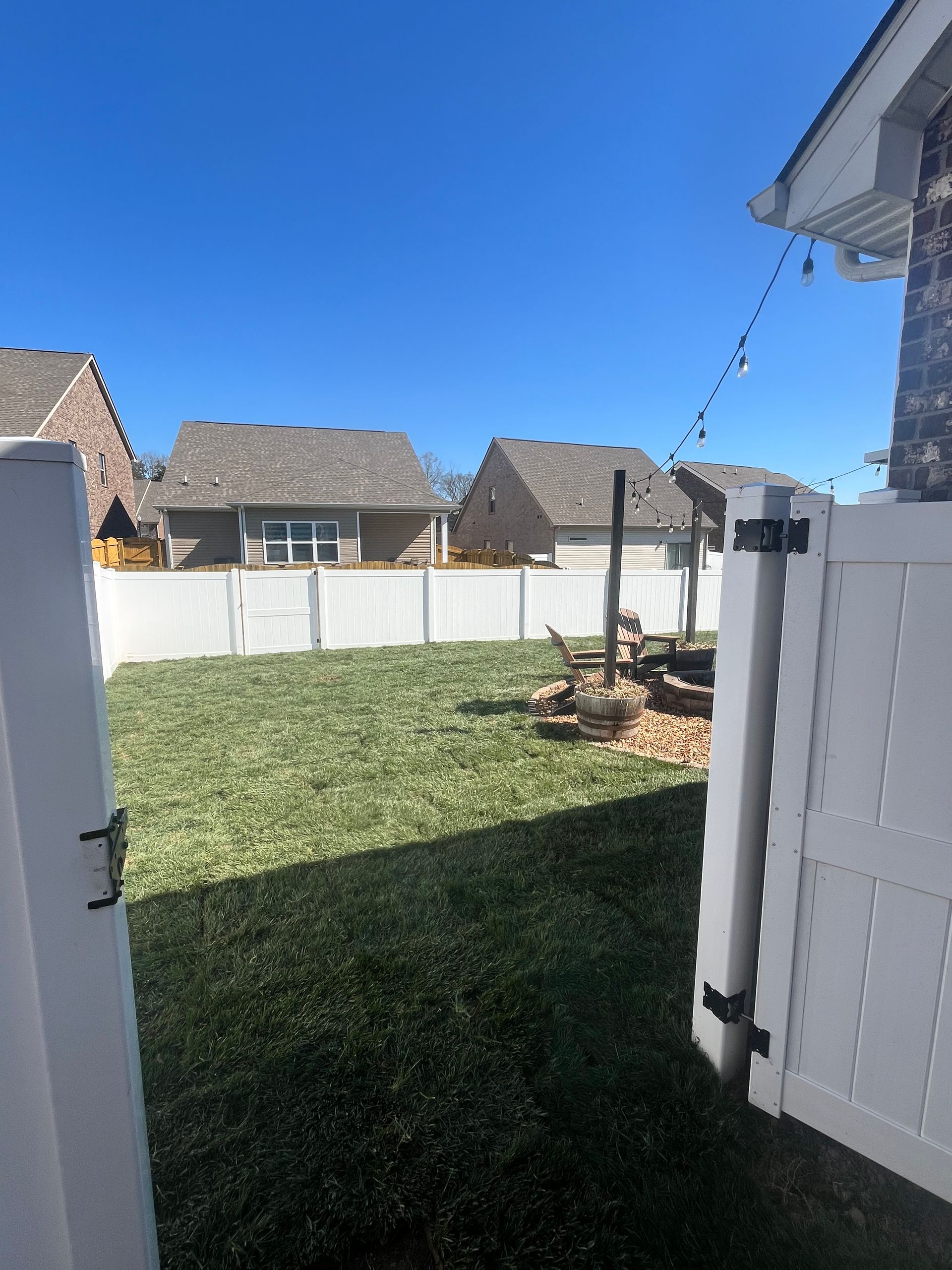 White fence opens to a green yard, fire pit, and neighboring houses under a blue sky.