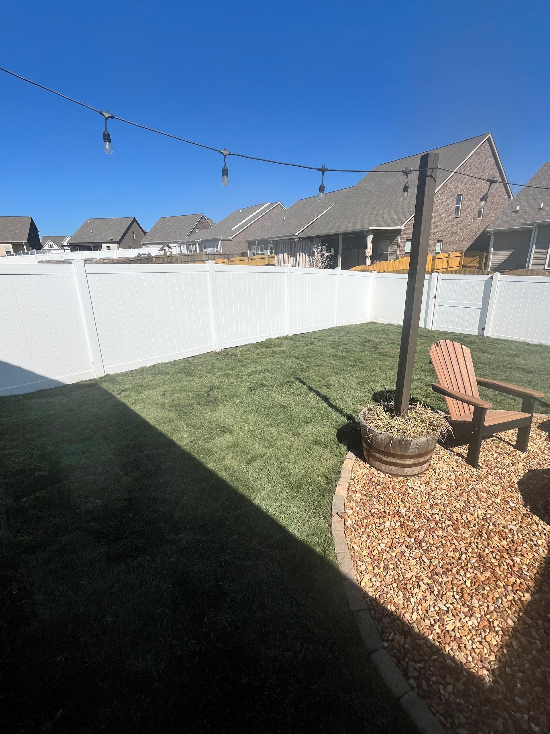Backyard with green grass, white fence, string lights, wooden chair, and blue sky.