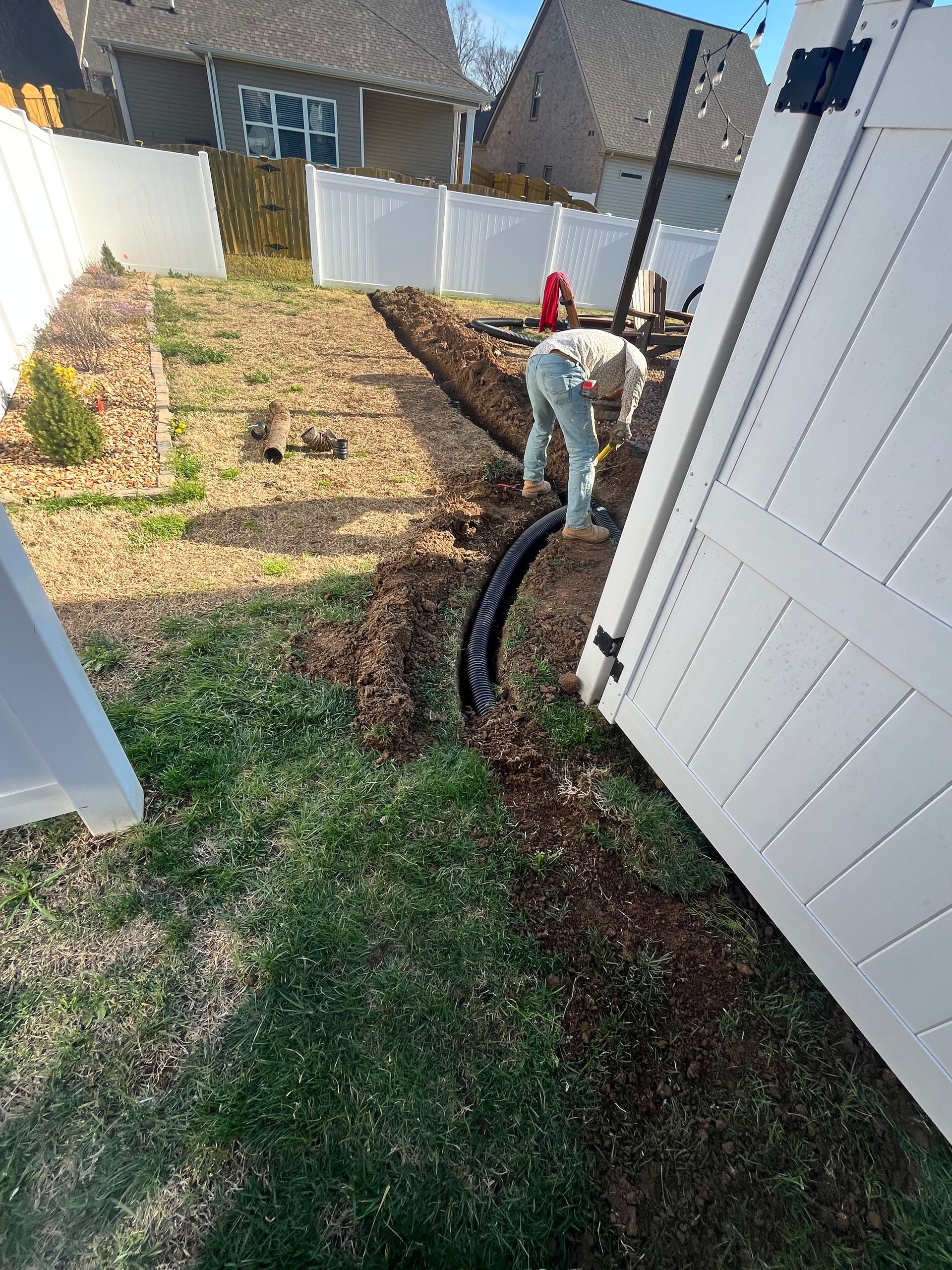 A person digs a trench in a backyard near a fence. A black drainage pipe is being installed.
