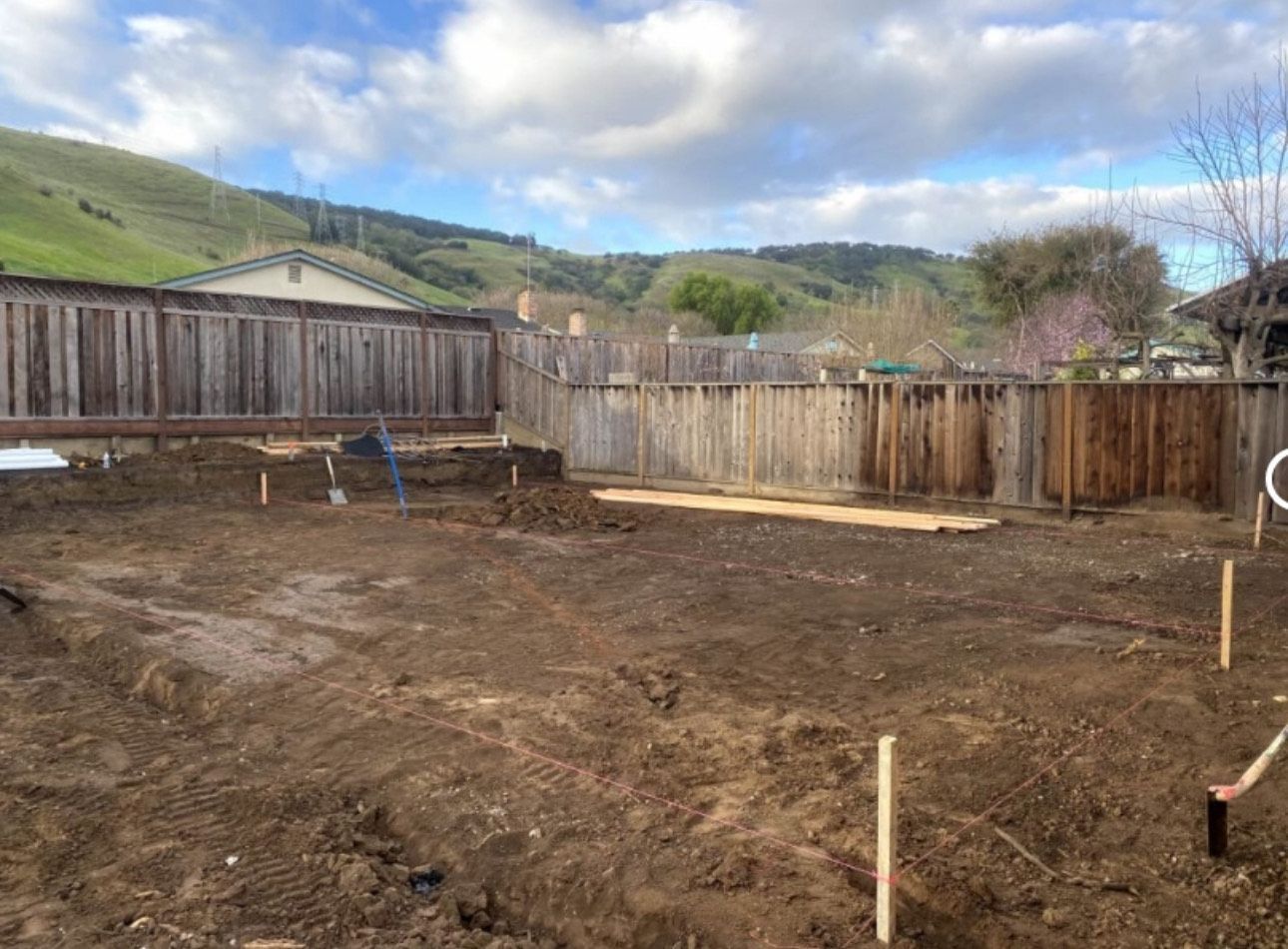 A backyard with a dirt patch and wooden fence against a hilly landscape under a partly cloudy sky.