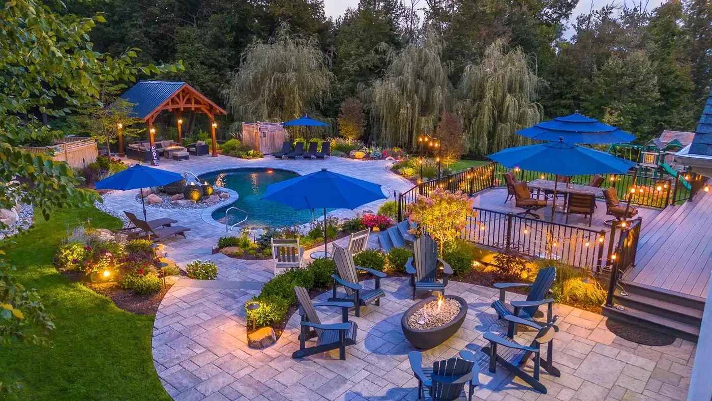 A landscaped backyard at twilight with a lit swimming pool, fire pit, stone patio, and blue patio umbrellas.