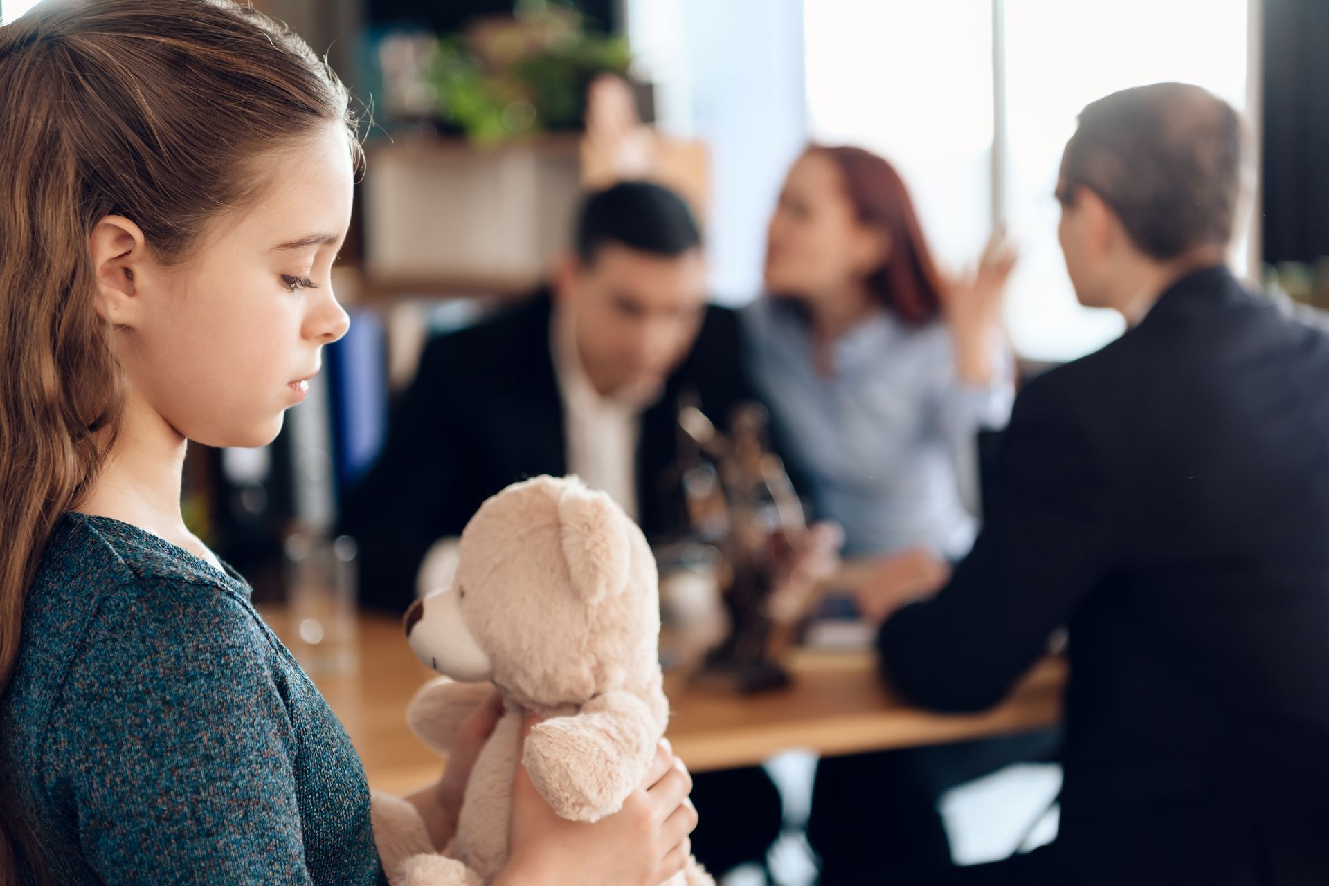 A sad girl with a teddy bear is standing while her parents argue in a lawyer’s office.