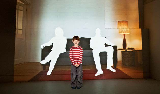 A boy is standing in front of a projection of two people sitting on a couch