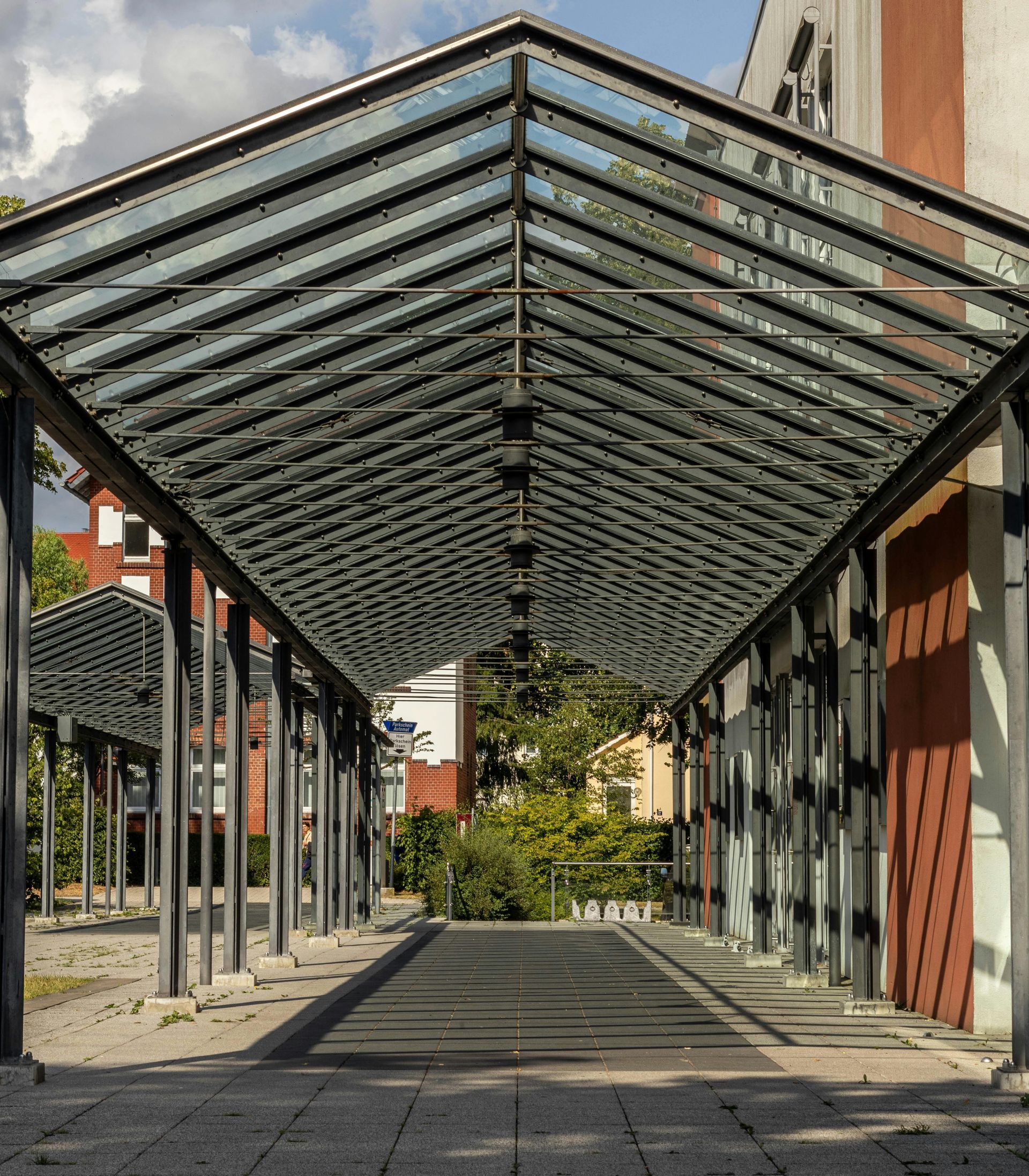 Covered walkway with a glass roof and metal columns leading toward trees and buildings