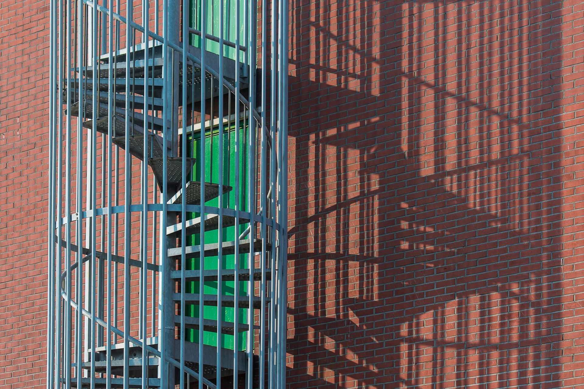 A spiral staircase behind metal bars, casting a shadow on a brick wall.