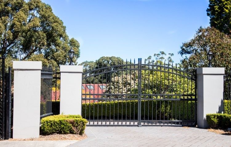 Black wrought-iron gate between white pillars at a sunny driveway entrance