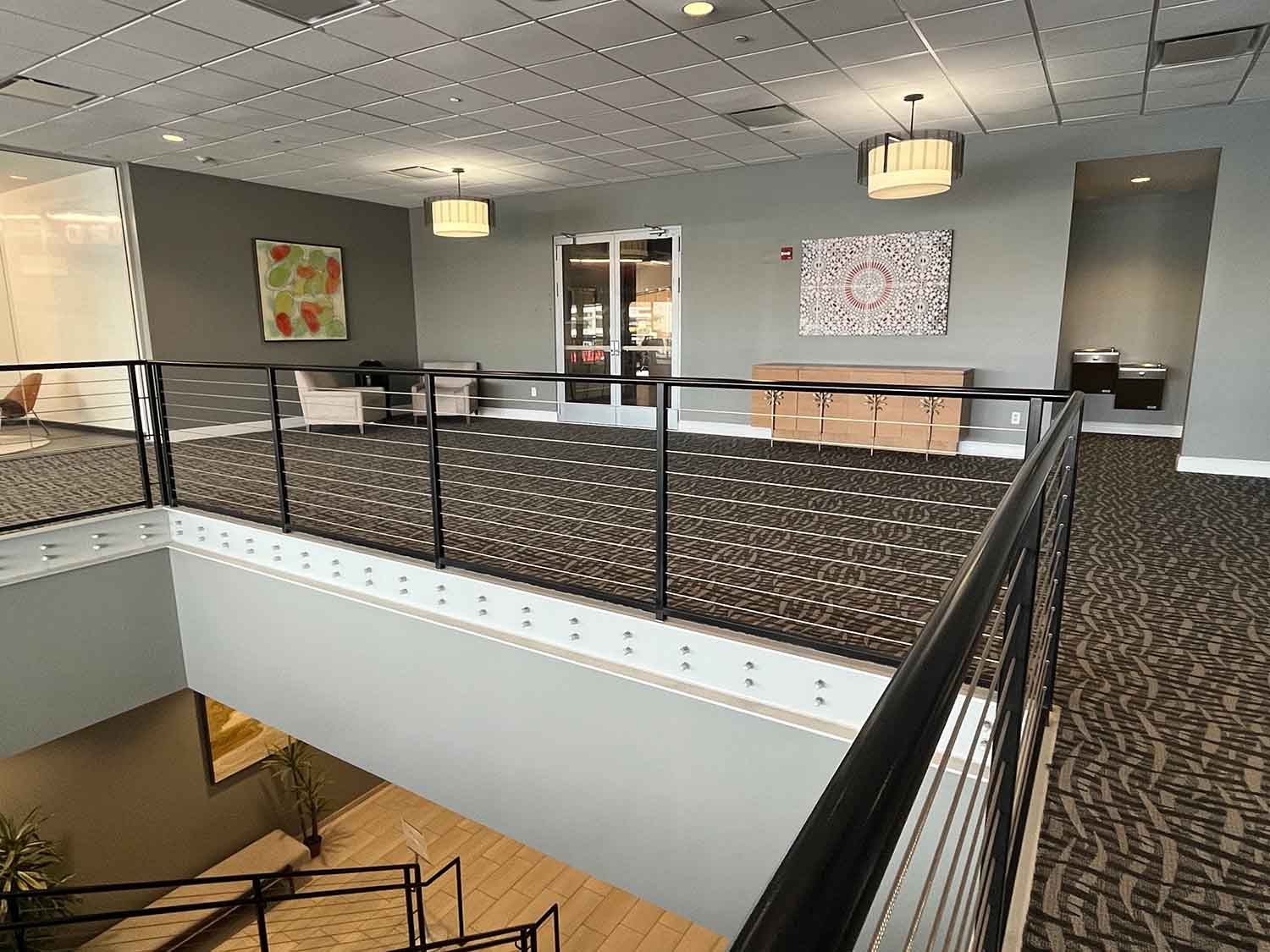 Office lobby with carpeted walkway, glass railing, and modern pendant lights over a stair opening