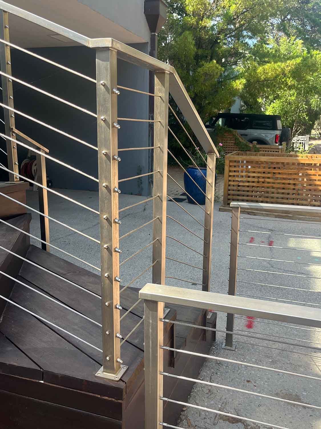 Outdoor deck with metal cable railing, stairs, and a driveway with parked cars in the background