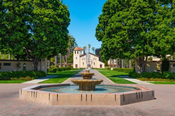 Fountain in a brick-lined walkway leading to a beige building flanked by large green trees.